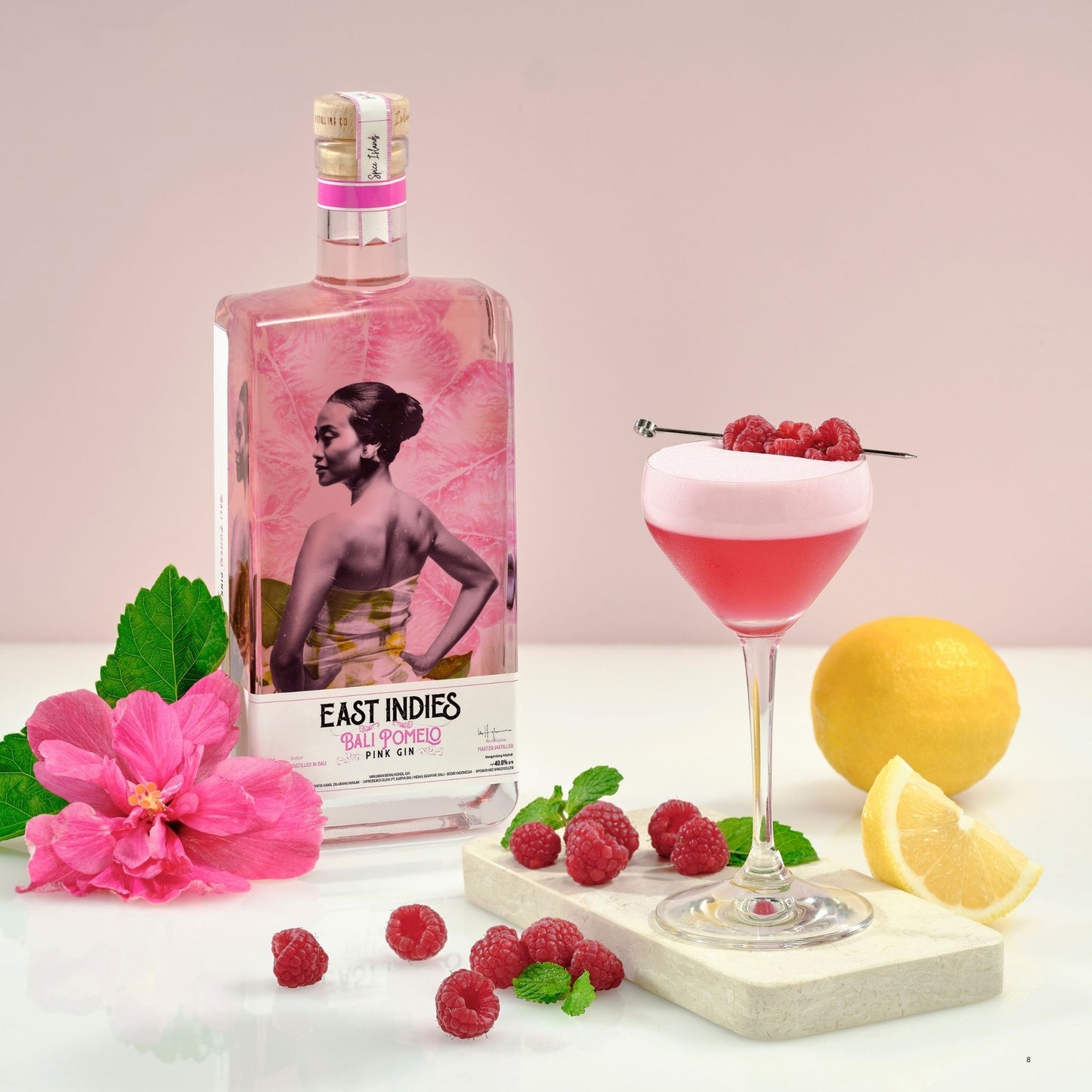 A bottle of East Indies Gin and Tonic cocktail mix alongside a prepared pink layered cocktail in a coupe glass with raspberries, set on a white surface with a pink background and styled with a lemon, flower, mint, and raspberries.