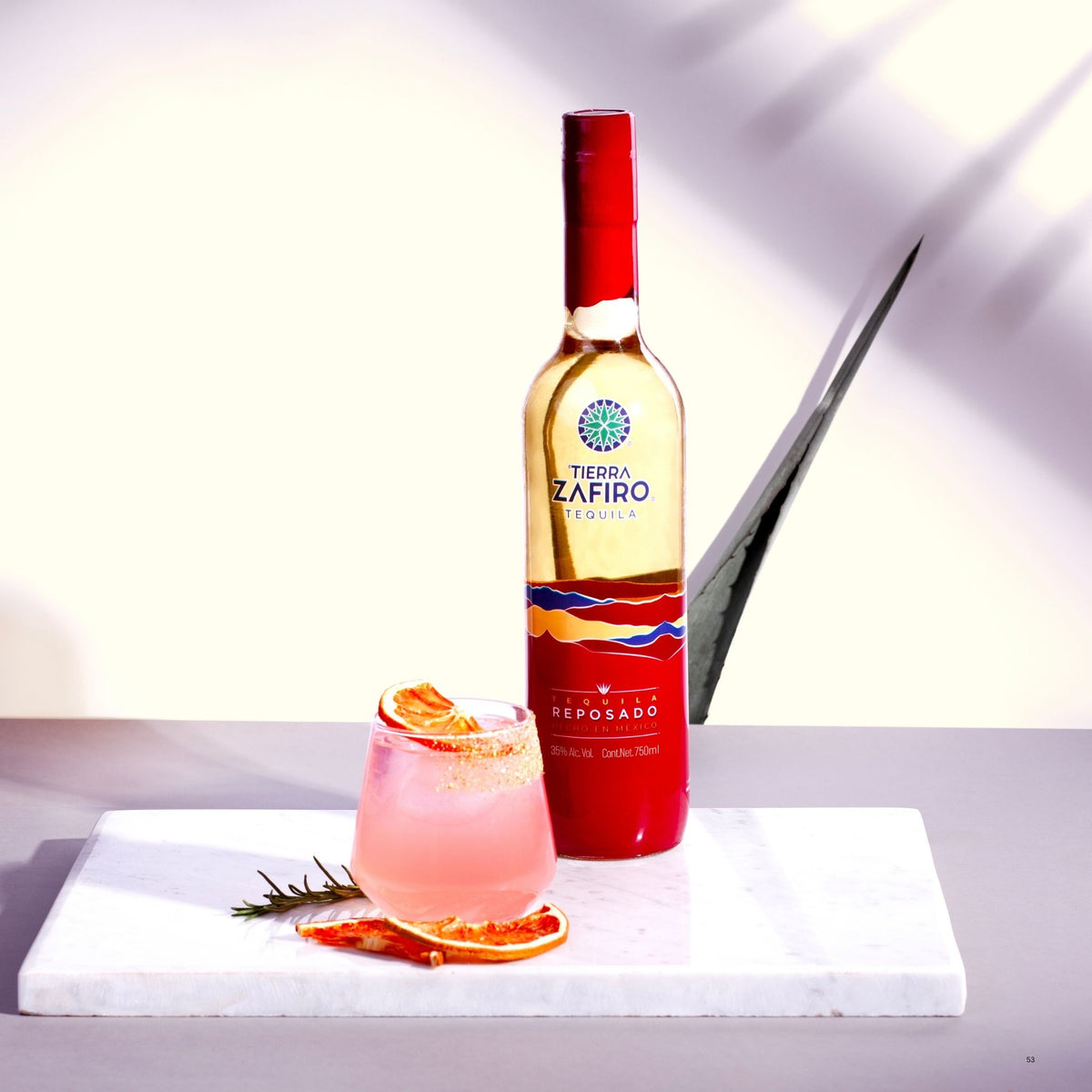 A bottle of Zafiro Reposado tequila stands on a white marble board next to a pink cocktail glass garnished with orange slices, with a rosemary sprig, against a clean white background with soft shadows.