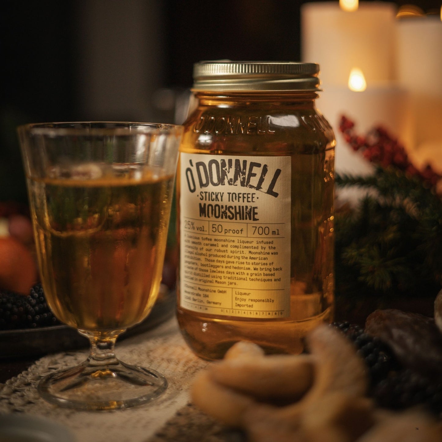 A glass of amber liquid sits beside a large clear jar labeled 'O'Donnell Sticky Toffee Moonshine' on a rustic tabletop with warm candlelight in the background.