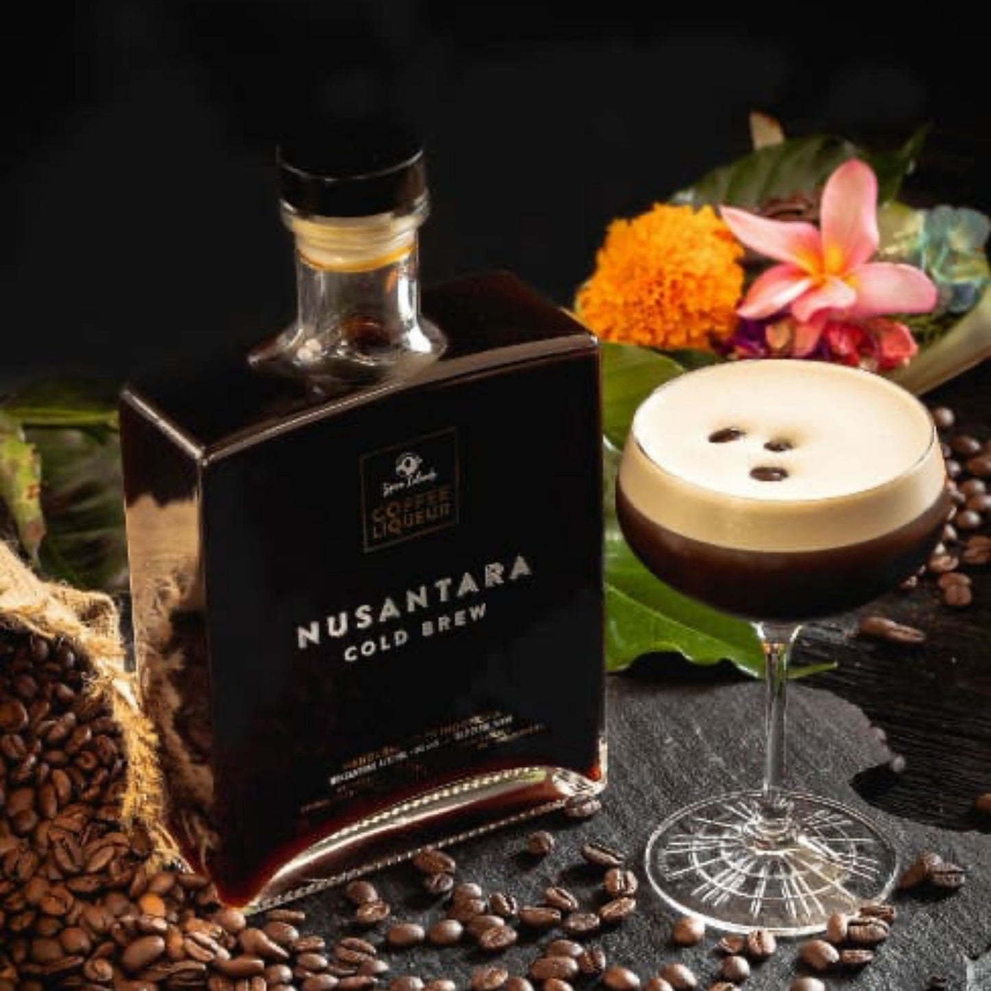 A bottle of Nusantara Cold Brew coffee next to a prepared cold brew cocktail in a stemmed glass, styled with coffee beans, green leaves, and tropical flowers on a dark background.