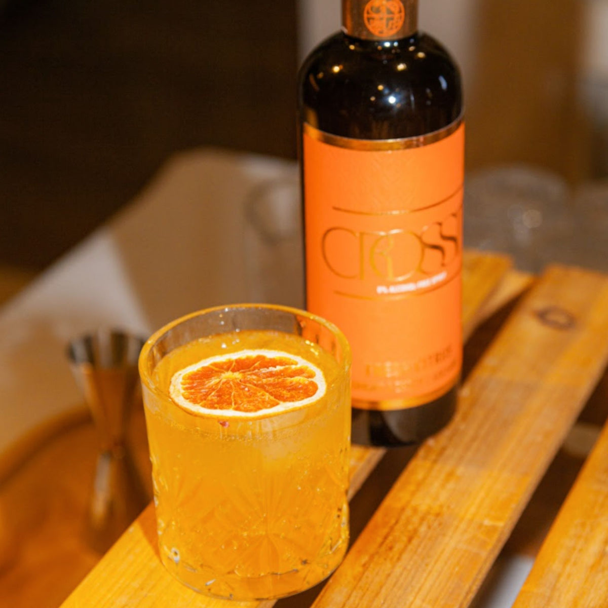 A glass of orange cocktail with a slice of orange on the rim, set on a wooden table next to a bottle with a bright orange label.