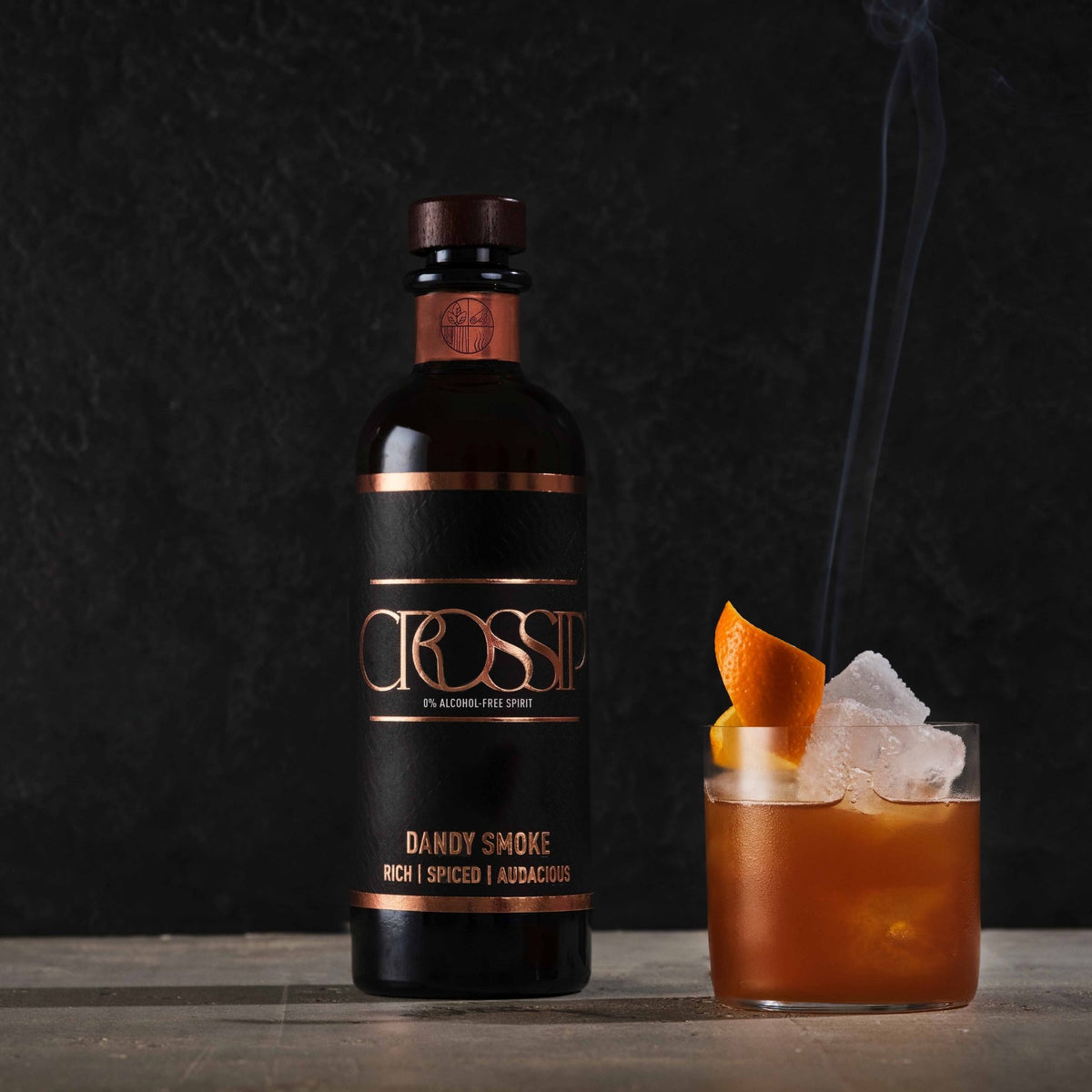 A bottle of Crossip Dandy Smoke alcoholic free spirit stands next to a rocks glass filled with an amber cocktail, ice, and an orange peel garnish, against a dark textured background.
