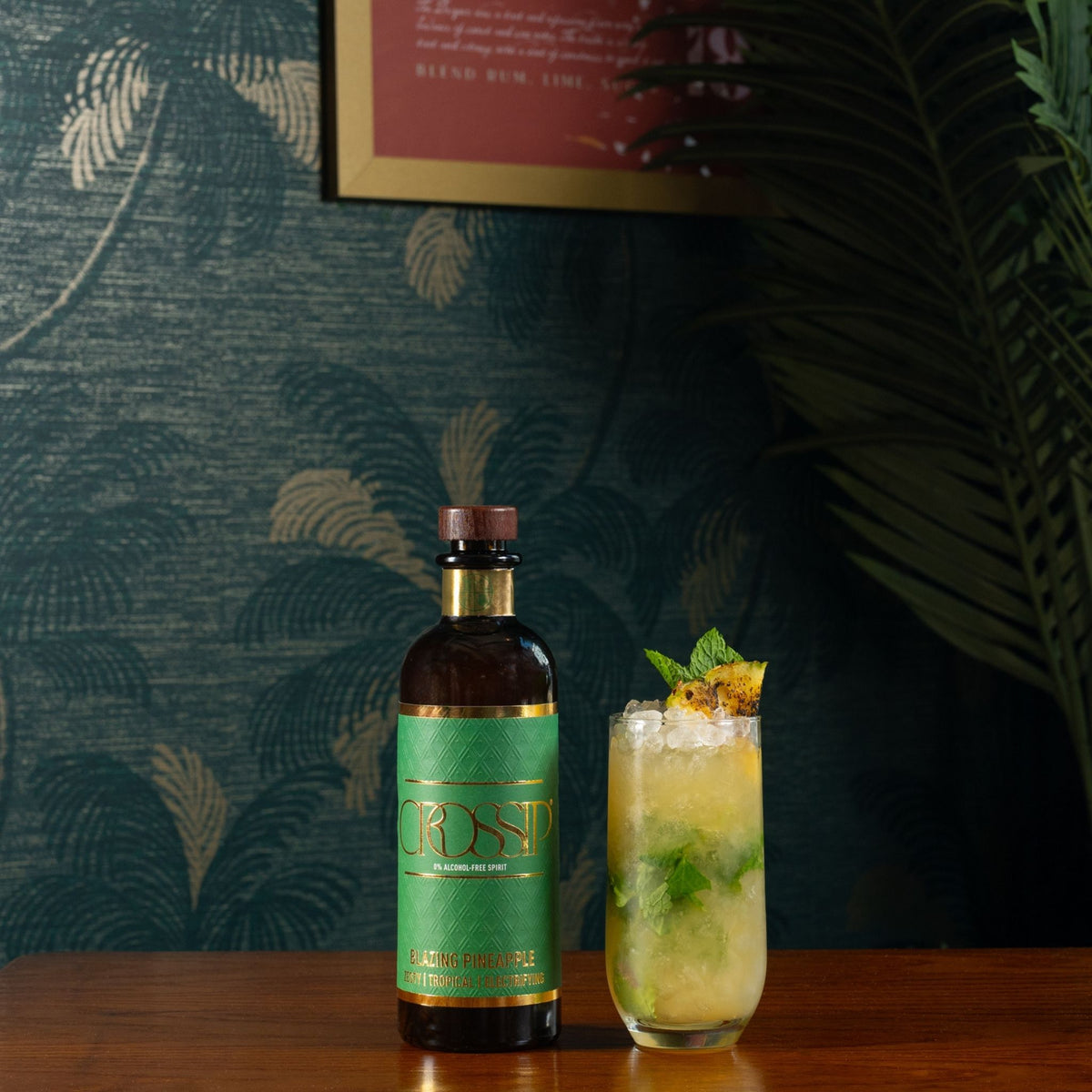 A bottle of CROSSIP Blazing Pineapple stands on a wooden table next to a tall glass filled with a pale yellow cocktail, garnished with mint and a citrus slice, set against a dark patterned wallpaper background.