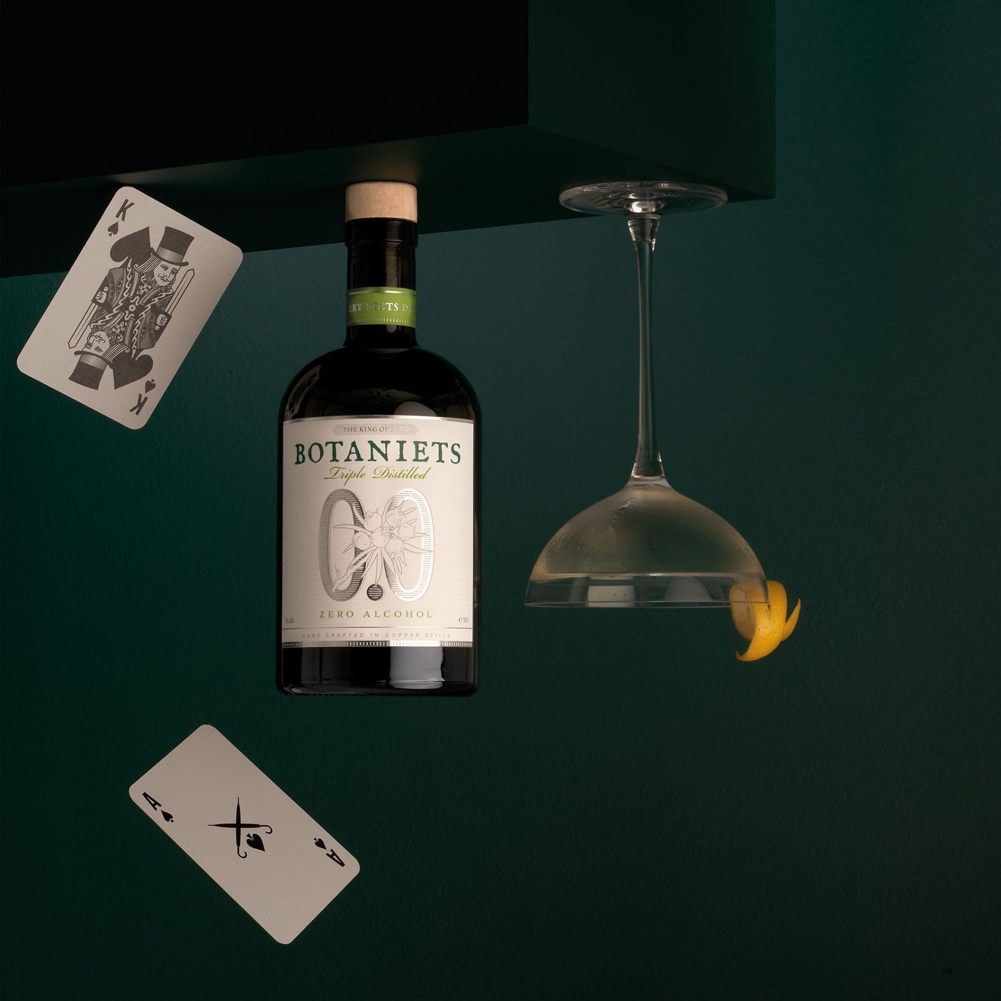 A bottle of Botaniets Triple Distilled is suspended against a dark green background, surrounded by floating playing cards and an upside-down wine glass with a yellow flower resting in the bowl. The brand name 'Botaniets' is clearly visible on the bottle label.
