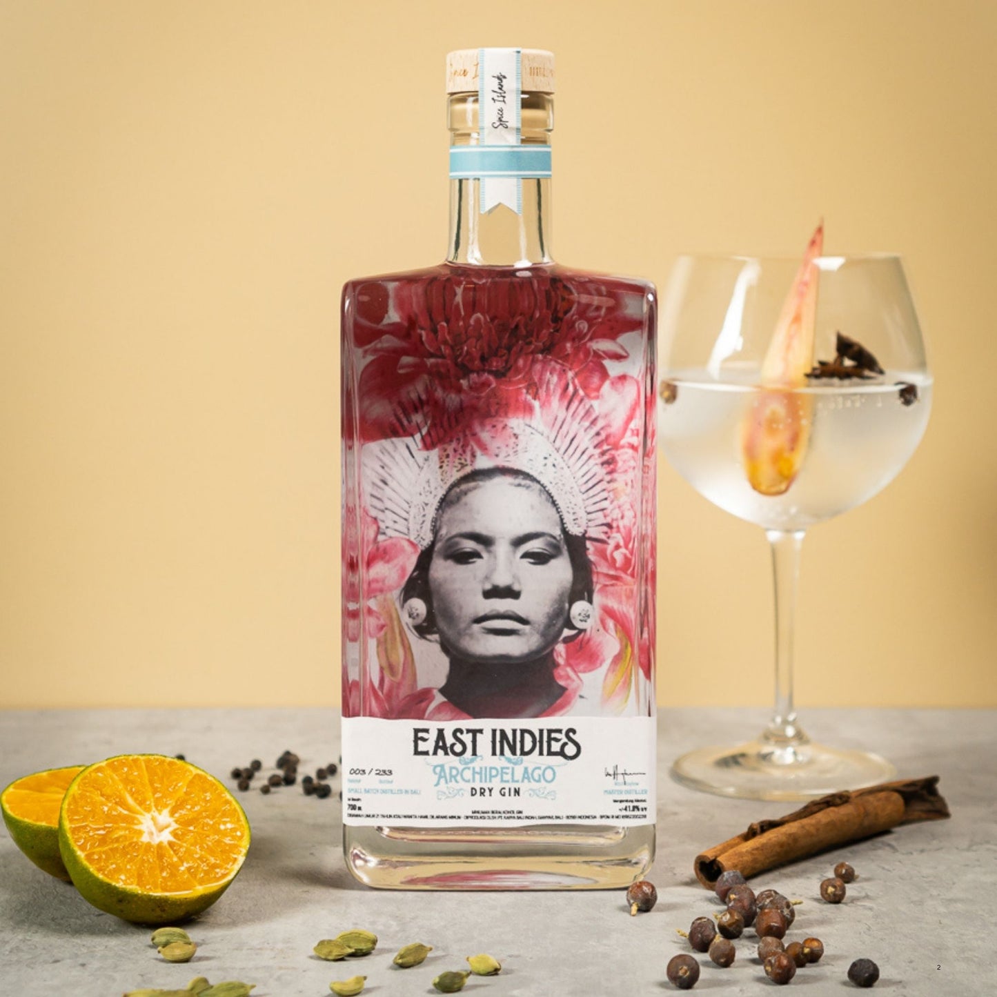 A bottle of East Indies Archipelago 250ml Club rum with a colorful label, styled on a gray surface with an orange slice, cinnamon stick, cloves, and a glass of clear cocktail against a beige background.