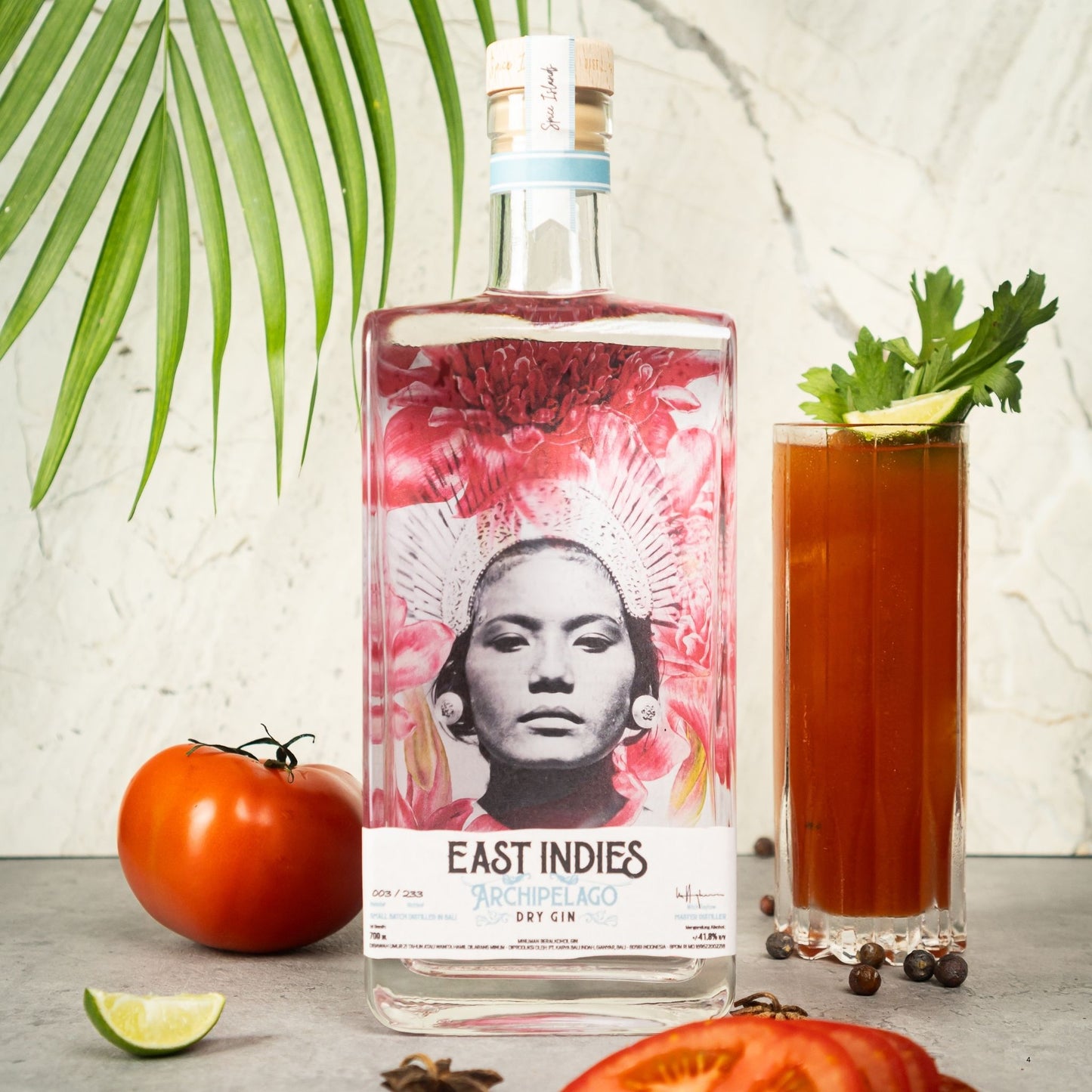 A bottle of East Indies Archipelago soy gin stands on a tabletop beside a tall glass of reddish-brown gin, garnished with cilantro and lime, with tomatoes, lime wedges, peppercorns, and a green palm leaf against a light stone wall.