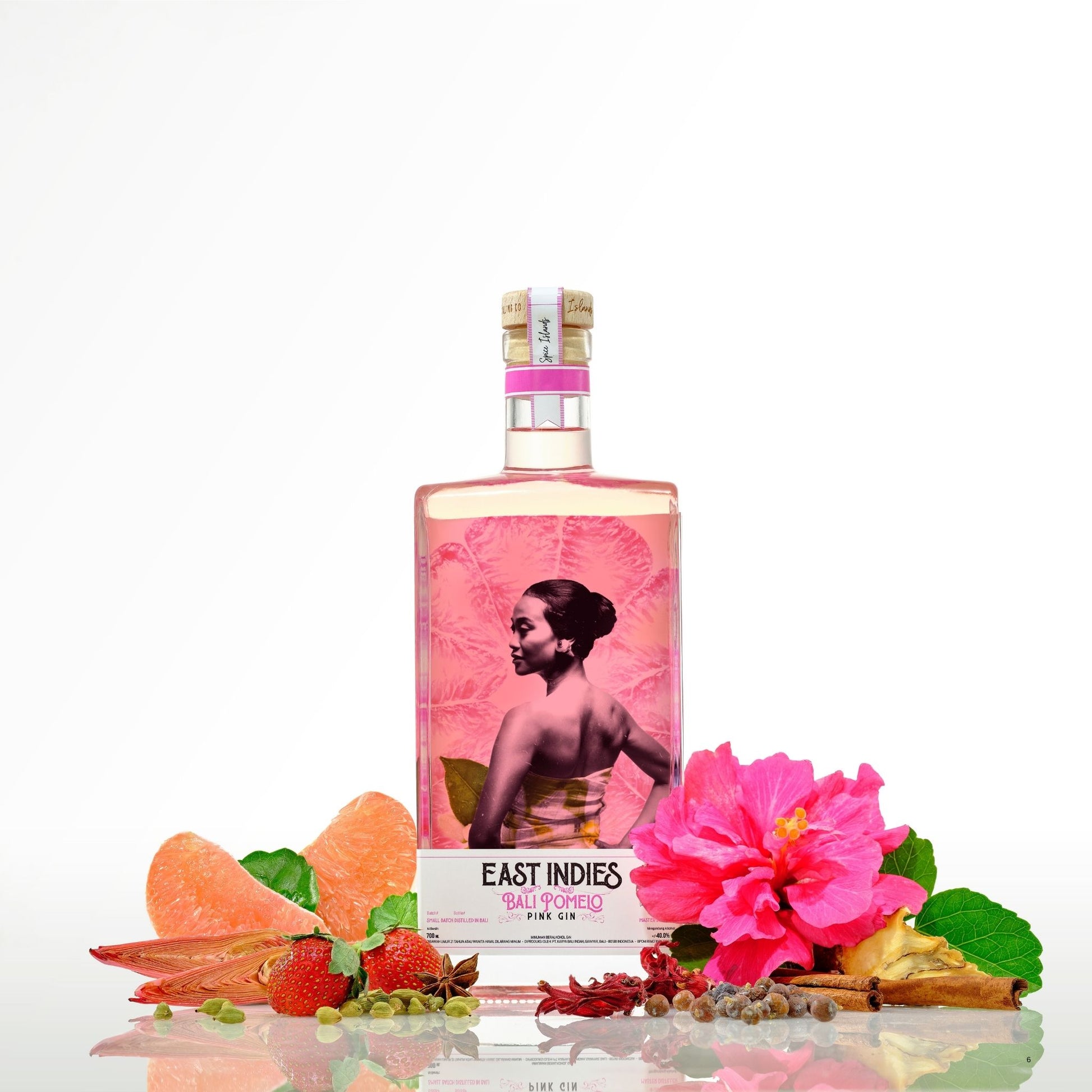 A bottle of East Indies Salted Gin with a pink citrus and floral label design, styled with pink hibiscus flowers, strawberries, orange slices, and spices on a reflective white surface against a white background.
