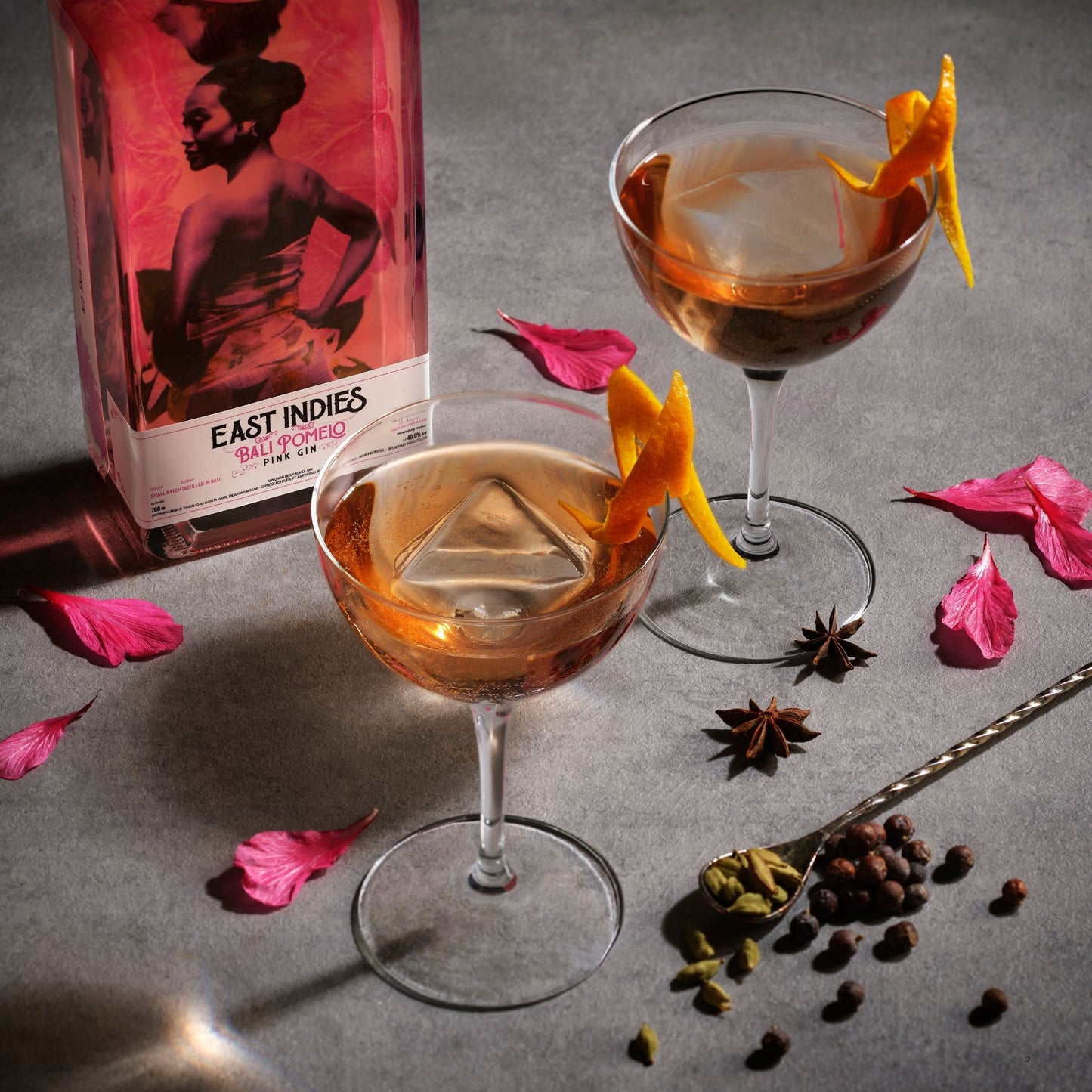 A bottle of East Indies Spiced Rum Cocktail mix is shown beside two coupe glasses filled with an amber cocktail and garnished with orange peel, on a gray surface with pink petals, star anise, and spices.