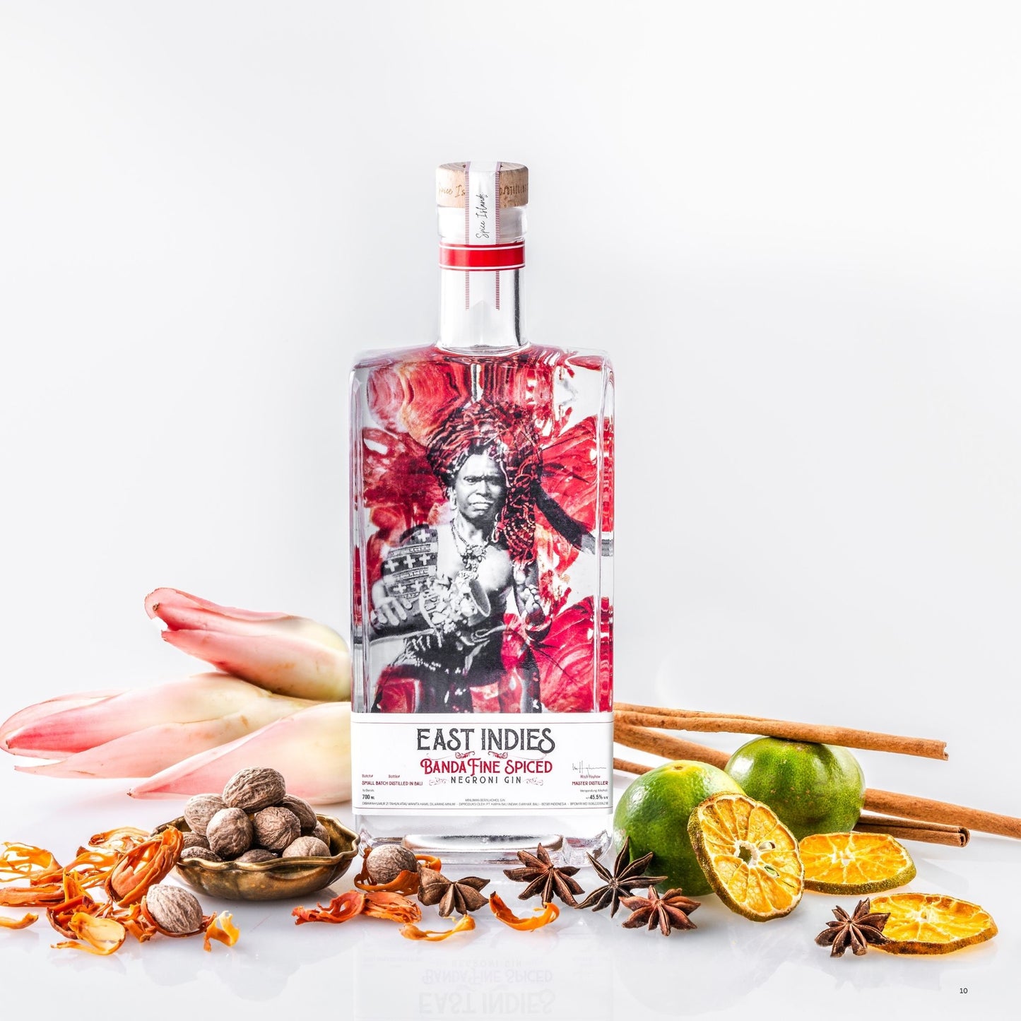 A bottle of East Indies Bangalore Spiced Gin with a colorful label, surrounded by botanical ingredients including pink tulips, cinnamon sticks, star anise, dried orange slices, and green and yellow citrus fruits on a white background.