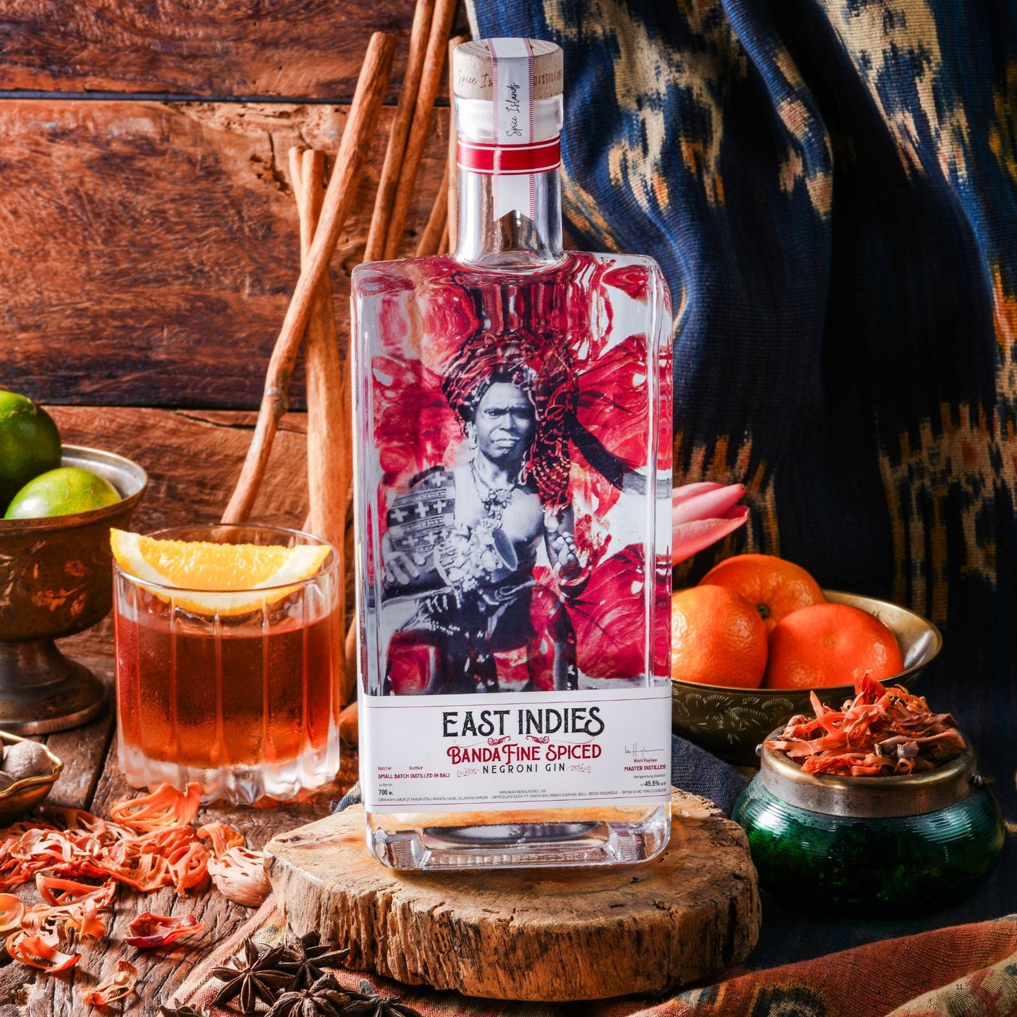 A bottle of East Indies Banga Fine Spiced Meethle Gin stands on a wooden slice in a styled bar-like setting with a filled cocktail glass, citrus fruits, spices, and a patterned textile in the background.