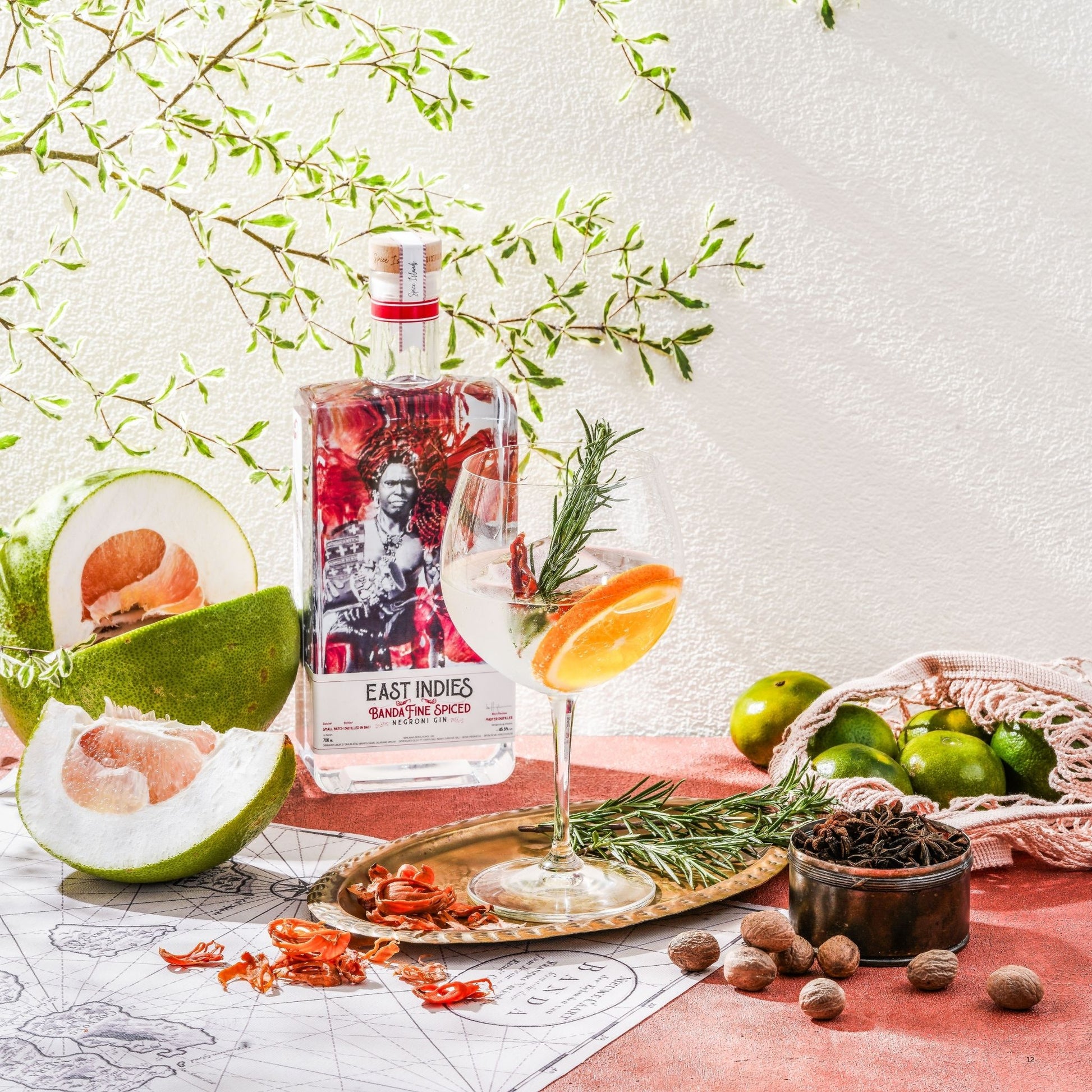 A clear glass bottle of East Indies Bombay Spirit Liqueur with a red-and-black illustrated label stands on a tabletop beside a filled coupe glass with an orange slice and rosemary garnish. The scene includes citrus fruit, spices, a basket, and leafy branches against a white wall, suggesting a styled product shot.