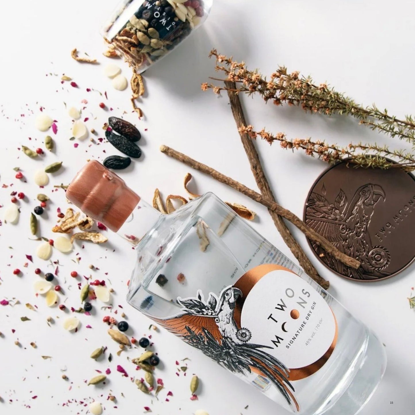 A clear glass bottle of Two Moths Condiment Oil lies on its side on a white surface, surrounded by loose spices, seeds, dried flowers, cinnamon sticks, and a bronze coin. The bottle has a white and copper label with a black bird illustration.