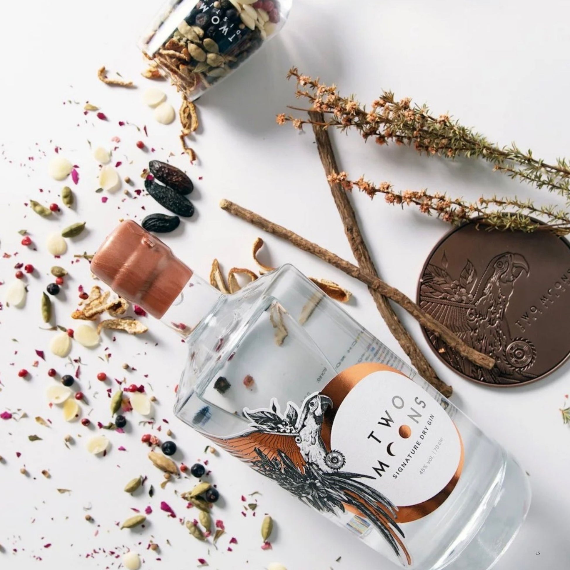 A clear glass bottle of Two Moths Condiment Oil lies on its side on a white surface, surrounded by loose spices, seeds, dried flowers, cinnamon sticks, and a bronze coin. The bottle has a white and copper label with a black bird illustration.
