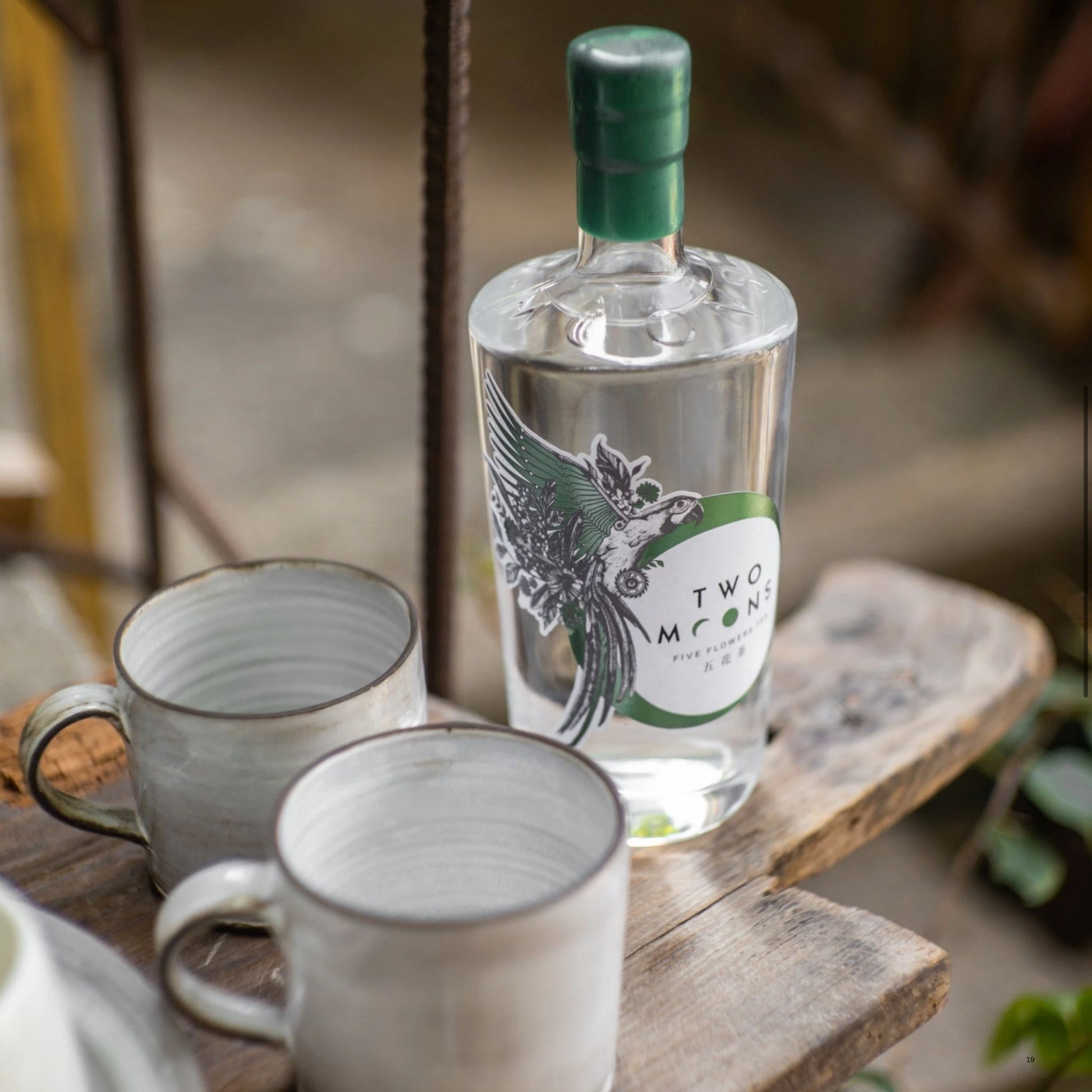 A clear glass bottle of Two Moon Gin with a green cap and a green-and-black bird label, placed on a rustic wooden board beside two white ceramic mugs in an outdoor setting.