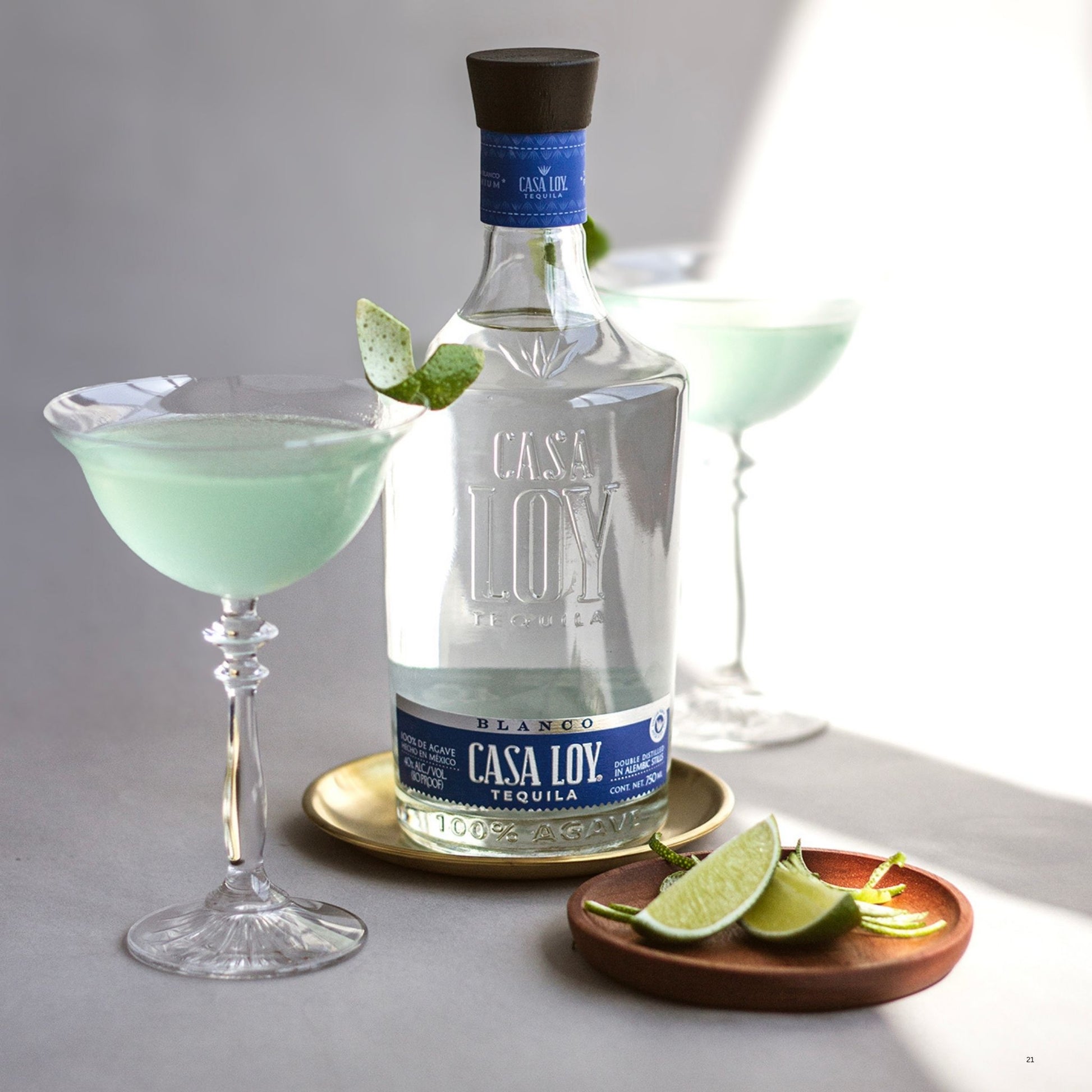 A bottle of Casa Loy tequila centered on a gray surface, flanked by two coupe glasses filled with a pale mint-green drink, plus lime wedges and a sprig on a small wooden dish.