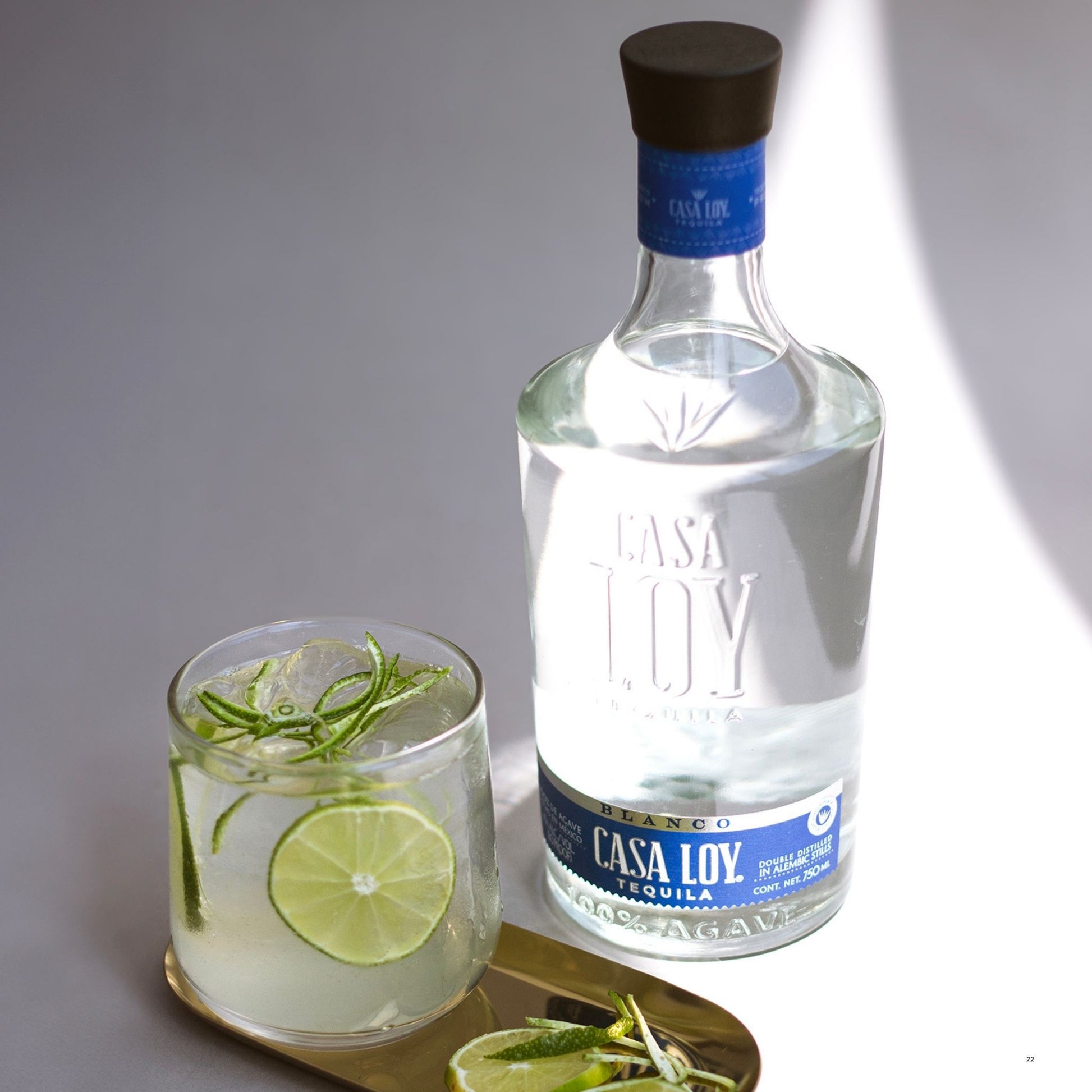 A bottle of Casa Loy Blanco Tequila stands next to a clear glass filled with a pale cocktail, garnished with lime slices and rosemary, on a small gold tray against a gray background.