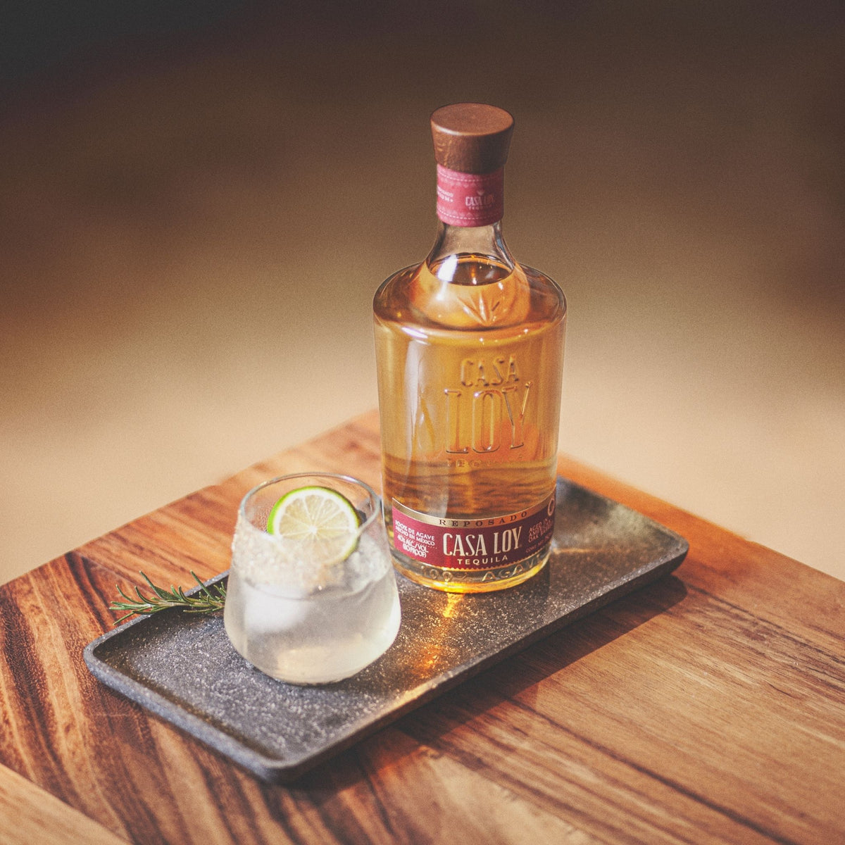A bottle of Casa Loni tequila and a glass of tequila-based cocktail on a dark rectangular tray, set on a wooden table with a warm, blurred background.
