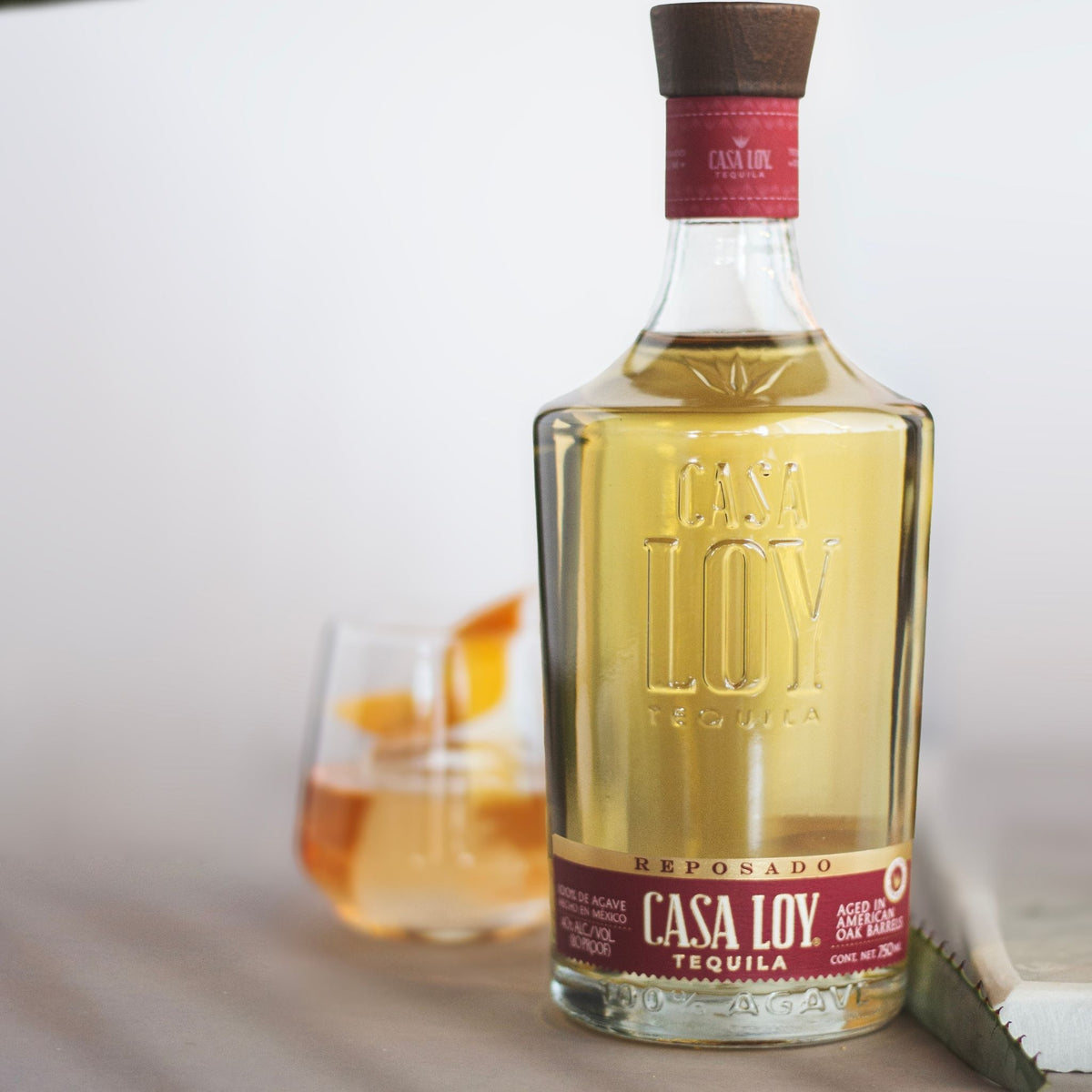 A bottle of Casa Loy Reposado Tequila with a clear glass body, golden-yellow liquid, and a dark brown neck wrap with a red label, set against a light background with a blurred glass of the same tequila and an orange slice in the background.
