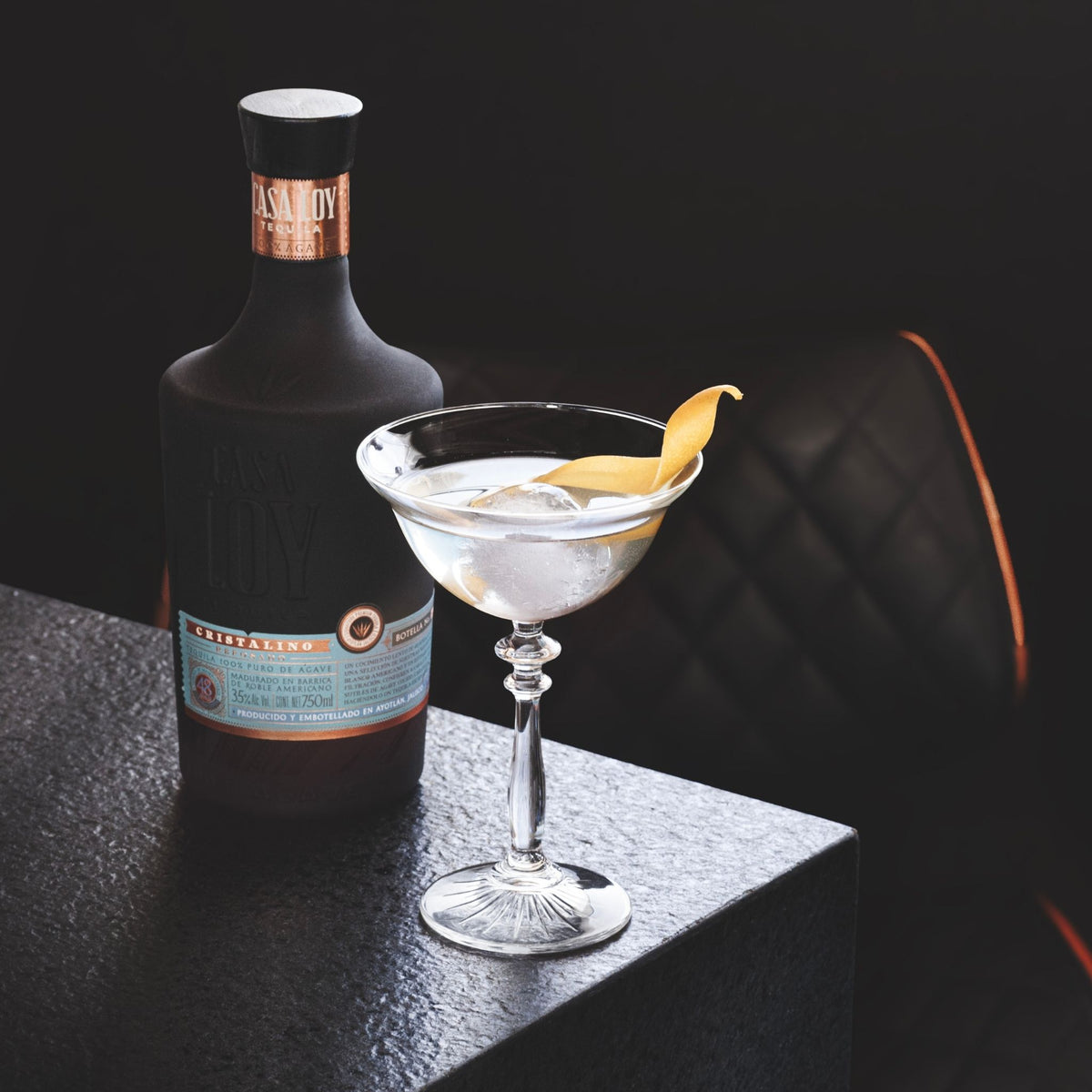 A bottle of Absolut London Gin and a filled martini glass with a citrus peel garnish, styled on a dark textured surface against a dark background.