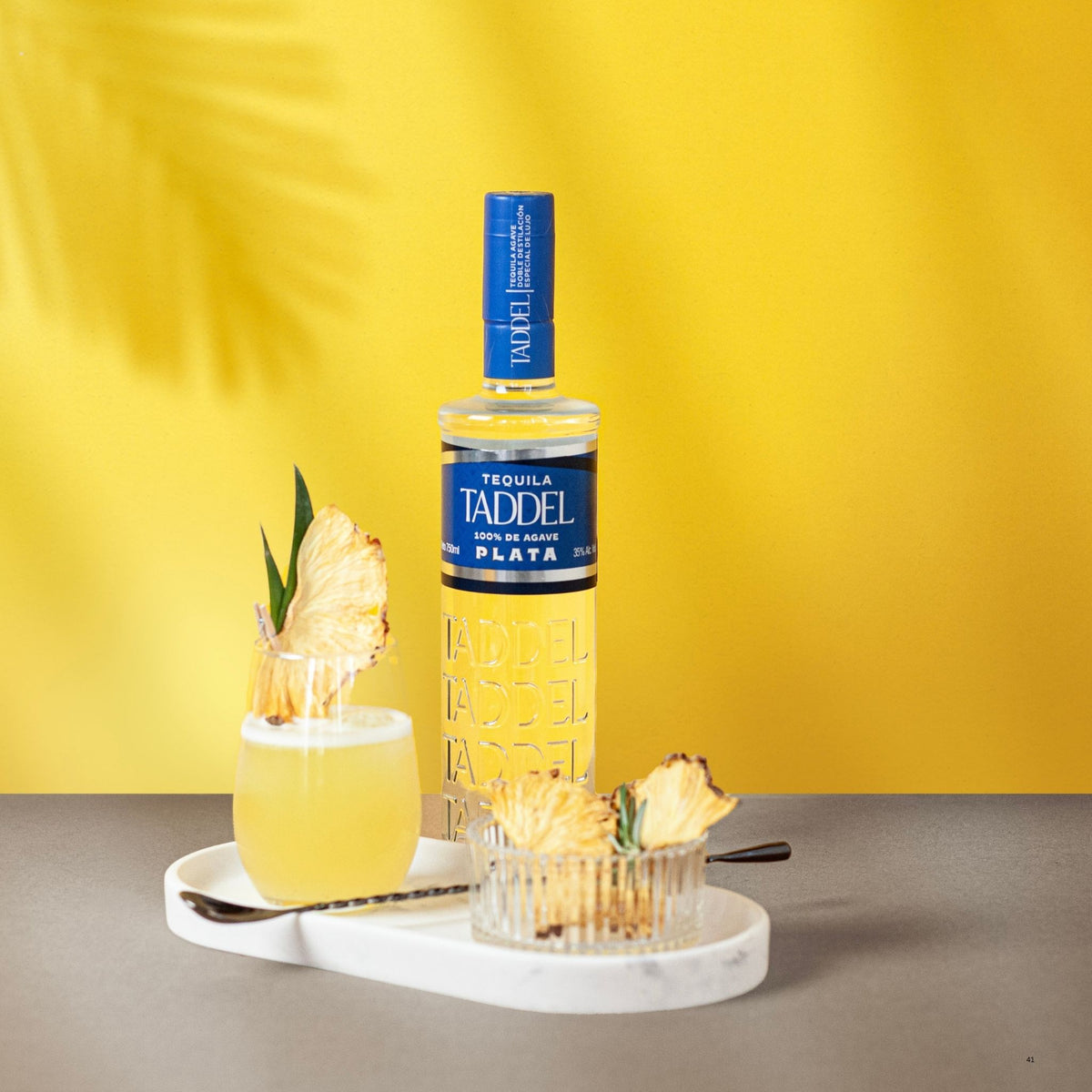A bottle of Taddel Plata tequila stands on a gray tabletop against a bright yellow wall, styled with a pineapple garnish and a glass of pale yellow drink on a white tray. A small bowl of pineapple slices sits beside it.