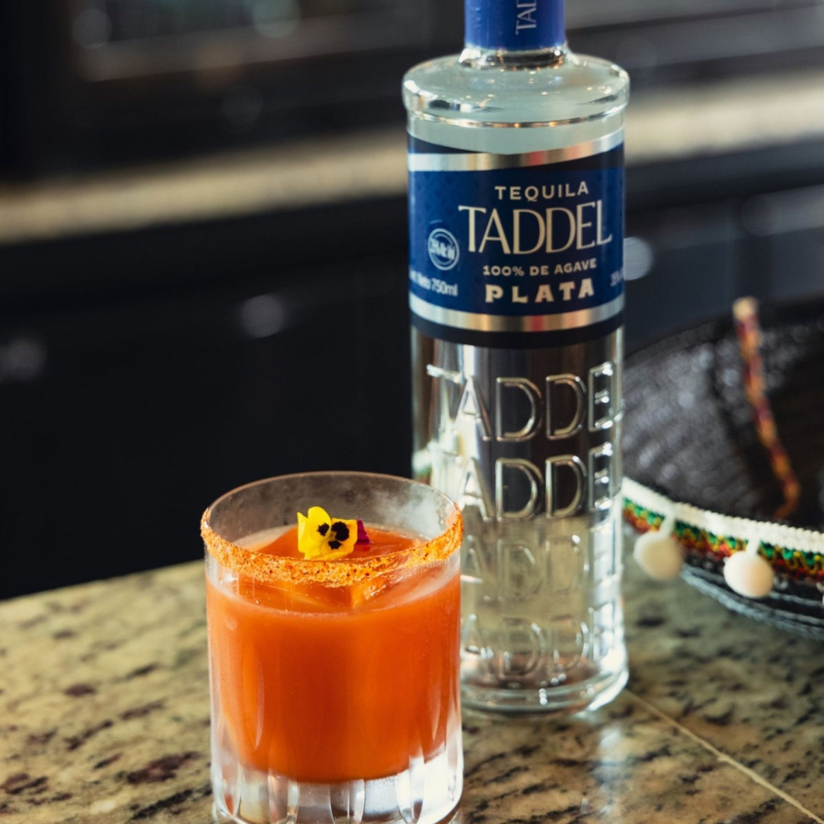A clear glass tumbler filled with an orange-red cocktail sits on a speckled countertop beside a large bottle of Tadeel Plata tequila. The glass rim is coated with reddish spice, and a small yellow flower garnish rests on top.