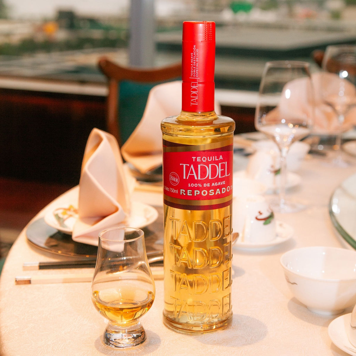 A bottle of Taddle Reposado tequila stands on a dining table beside a filled tequila glass, with wine glasses, folded napkins, and a softly blurred restaurant or bar background.