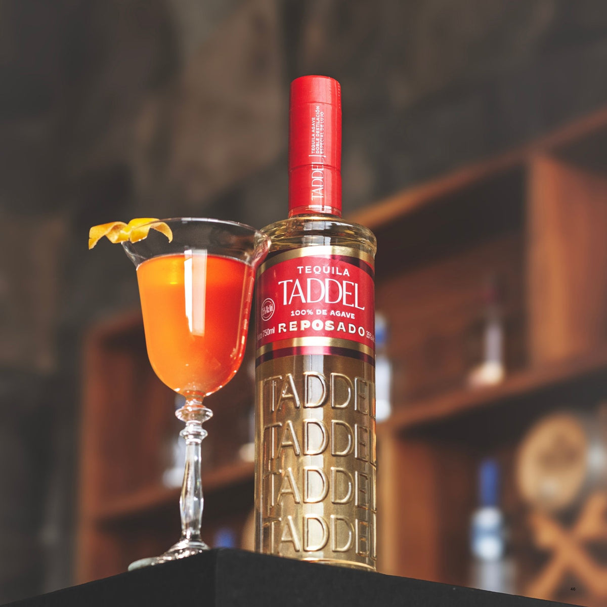A bottle of Tadeel Reposado tequila stands next to a filled coupe glass with an orange cocktail and a citrus garnish, set against a blurred bar background.