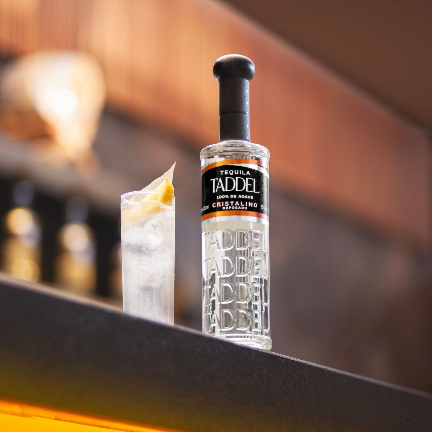 A bottle of Taddel Tequila 100% de Ancho Tequila Cristalino with a black stopper stands on a dark bar counter next to a clear highball glass filled with ice and a lemon wedge, against a warm, blurred bar background.