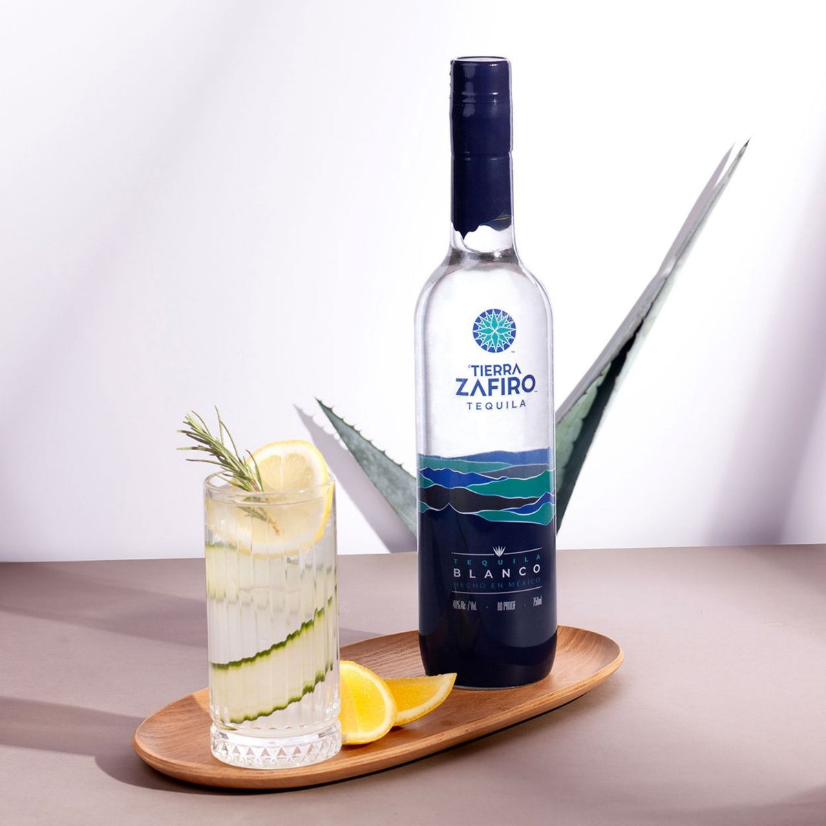 A bottle of Tierra Zafiro Tequila Blanco stands on a wooden tray beside a clear glass filled with a pale drink, lemon slices, cucumber strips, and a rosemary sprig, against a clean studio background.