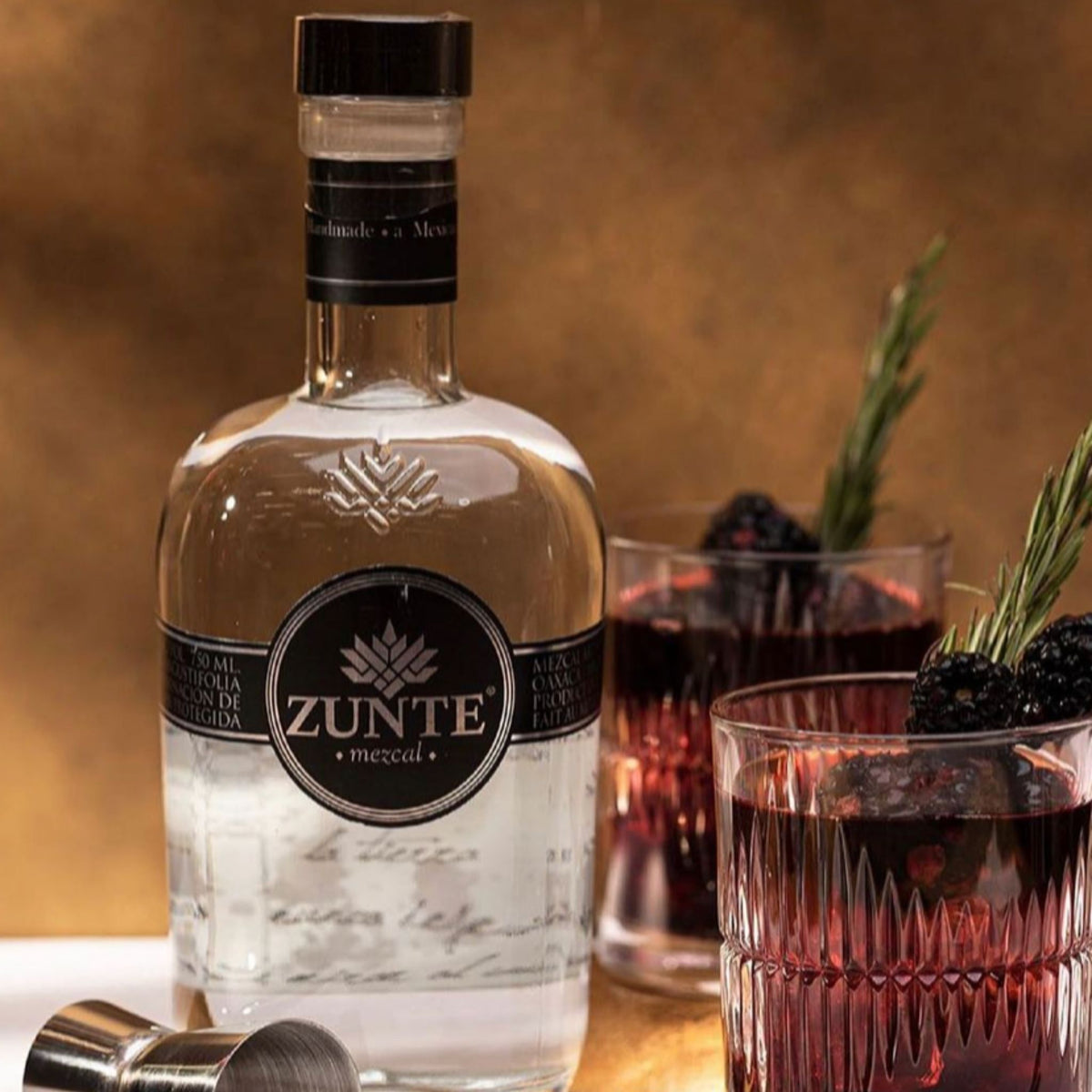 A bottle of Zunte Mezcal tequila with a black cap and black label, shown alongside two glasses of dark red cocktail and a metal jigger on a warm brown background.