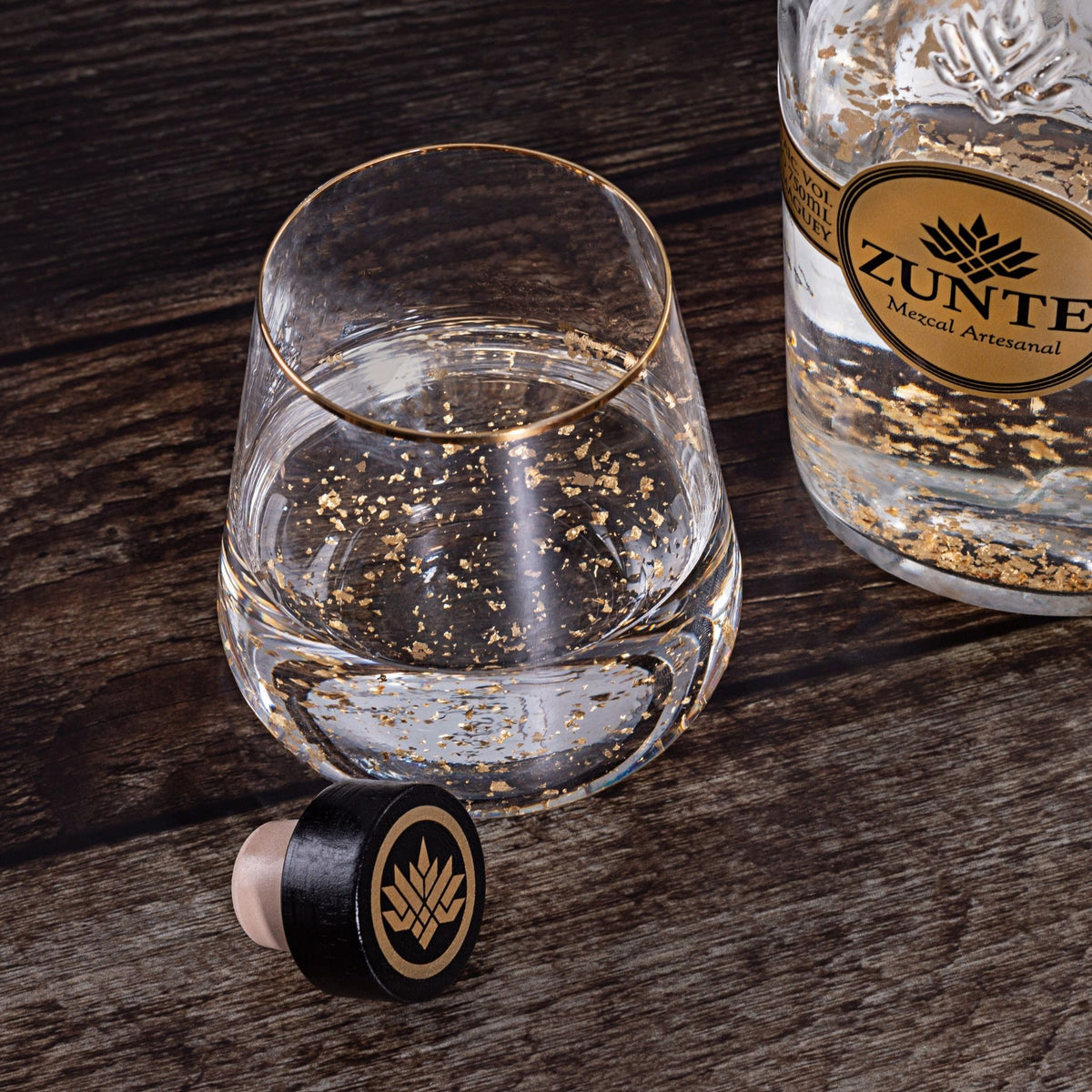 A clear glass tumbler with a gold rim and scattered gold flakes inside, shown on a dark wood surface beside a branded bottle and a black-and-gold bottle stopper. The bottle label reads ZUNTE.