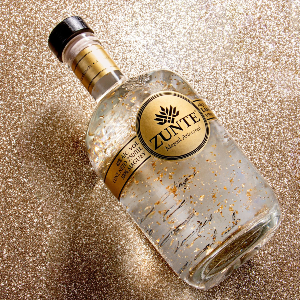 A bottle of ZUNTE Mezcal, decorated with gold flakes, lying diagonally on a sparkling gold glitter background.