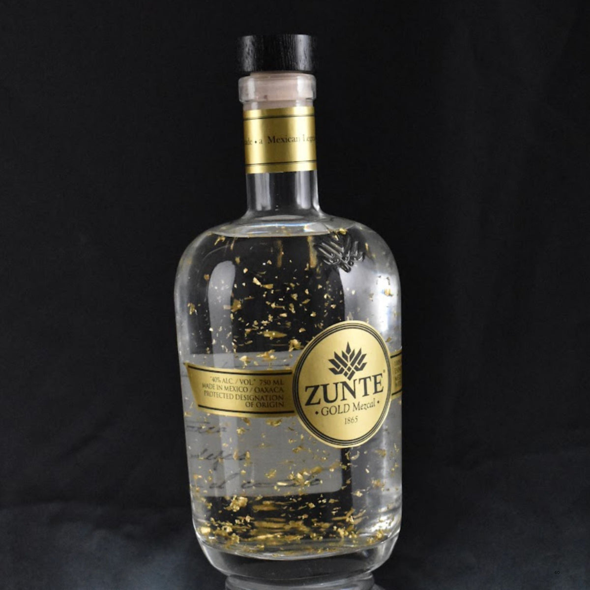 A clear glass bottle of Zunte Gold Mezcal, filled with shimmering gold flakes, set against a solid black background.