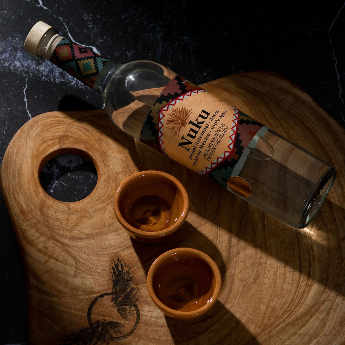 A clear glass bottle of Nuku tequila with a colorful geometric label, resting on a carved wooden serving board next to two small wooden shot glasses on a dark marble surface.