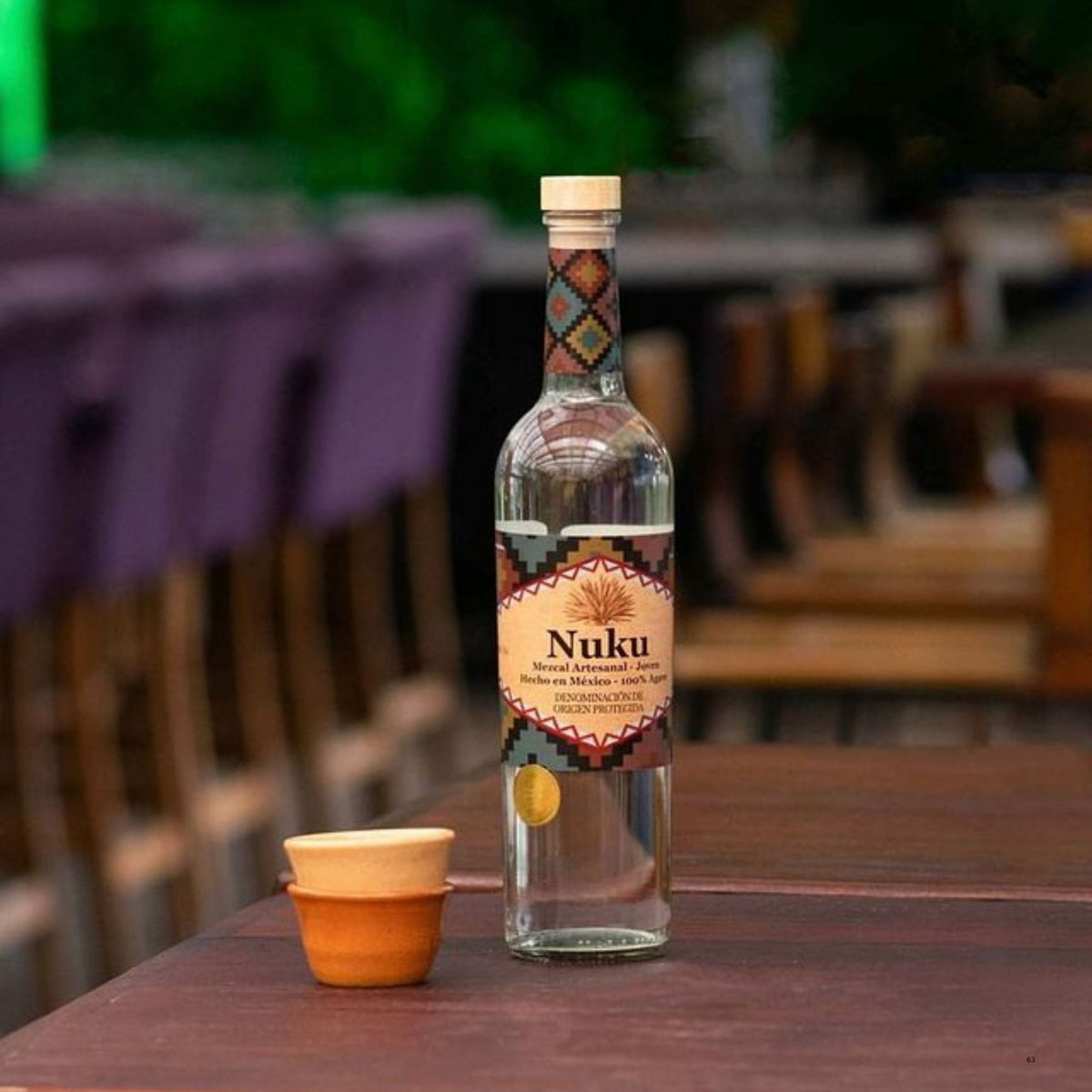 A clear glass bottle of Nuku mezcal on a wooden table next to a small orange and beige ceramic cup, with a blurred outdoor patio background.