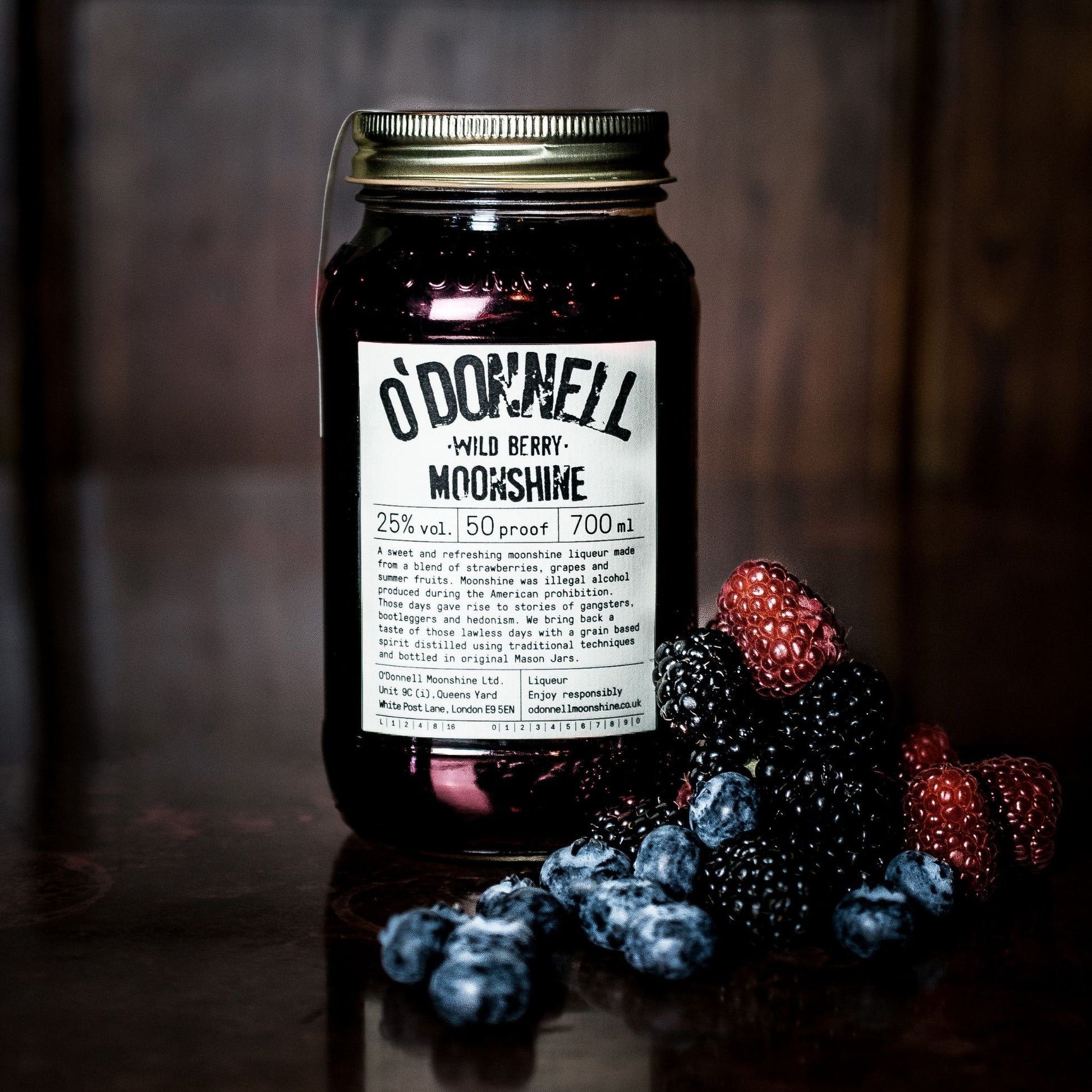 A bottle of O'Donnell Wild Berry Moonshine whiskey with a white label and gold cap, set on a dark reflective surface next to fresh blueberries, blackberries, and raspberries against a dark wooden background.