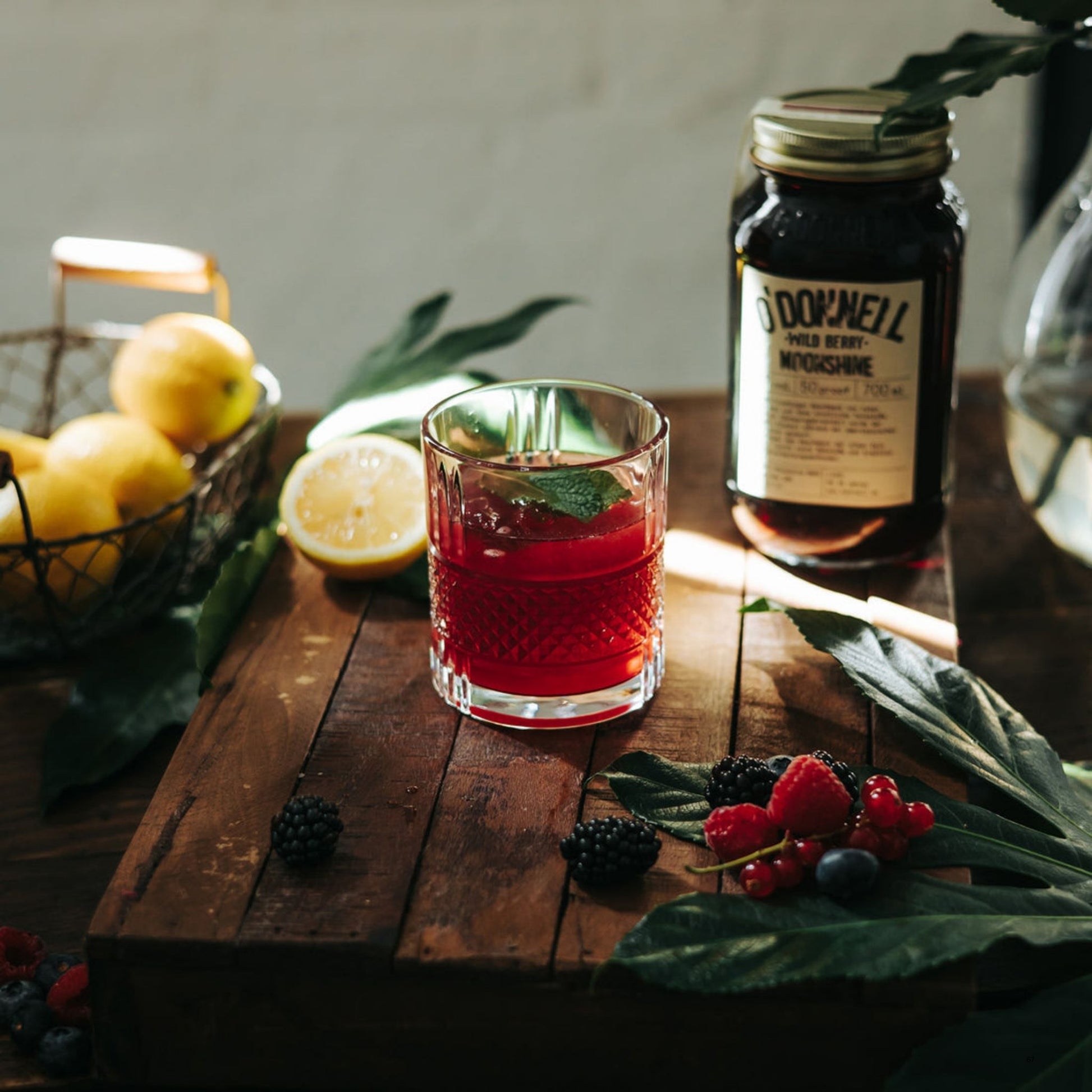 A styled tabletop scene with a clear tumbler glass filled with a dark red drink, a bottle of O’Donnell Moonshine.