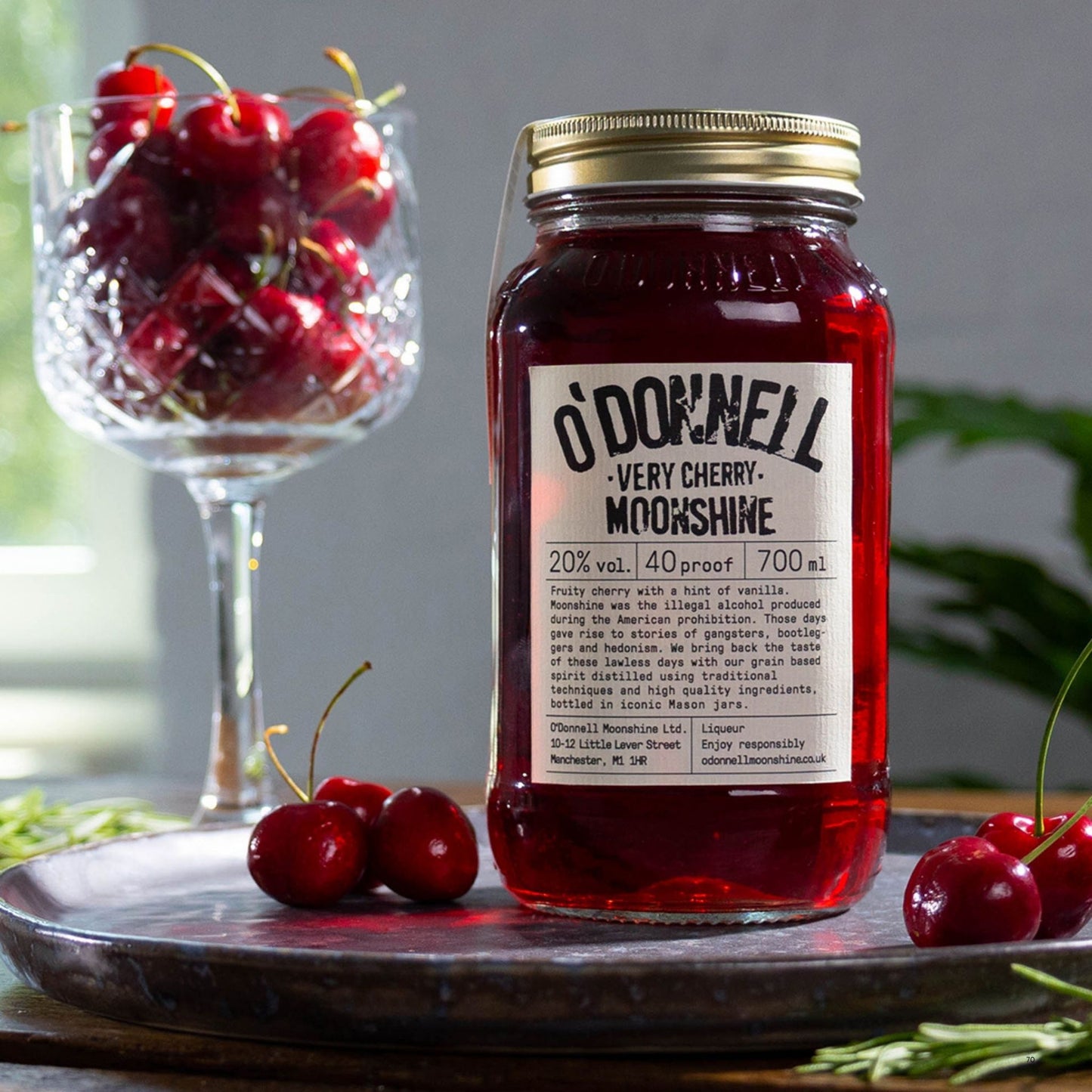 A glass jar of O'Donnell Very Cherry Moonshine liqueur with a gold lid and white label, placed on a dark tray with fresh cherries around it. A stemmed glass filled with cherries sits behind the jar against a softly blurred indoor background.