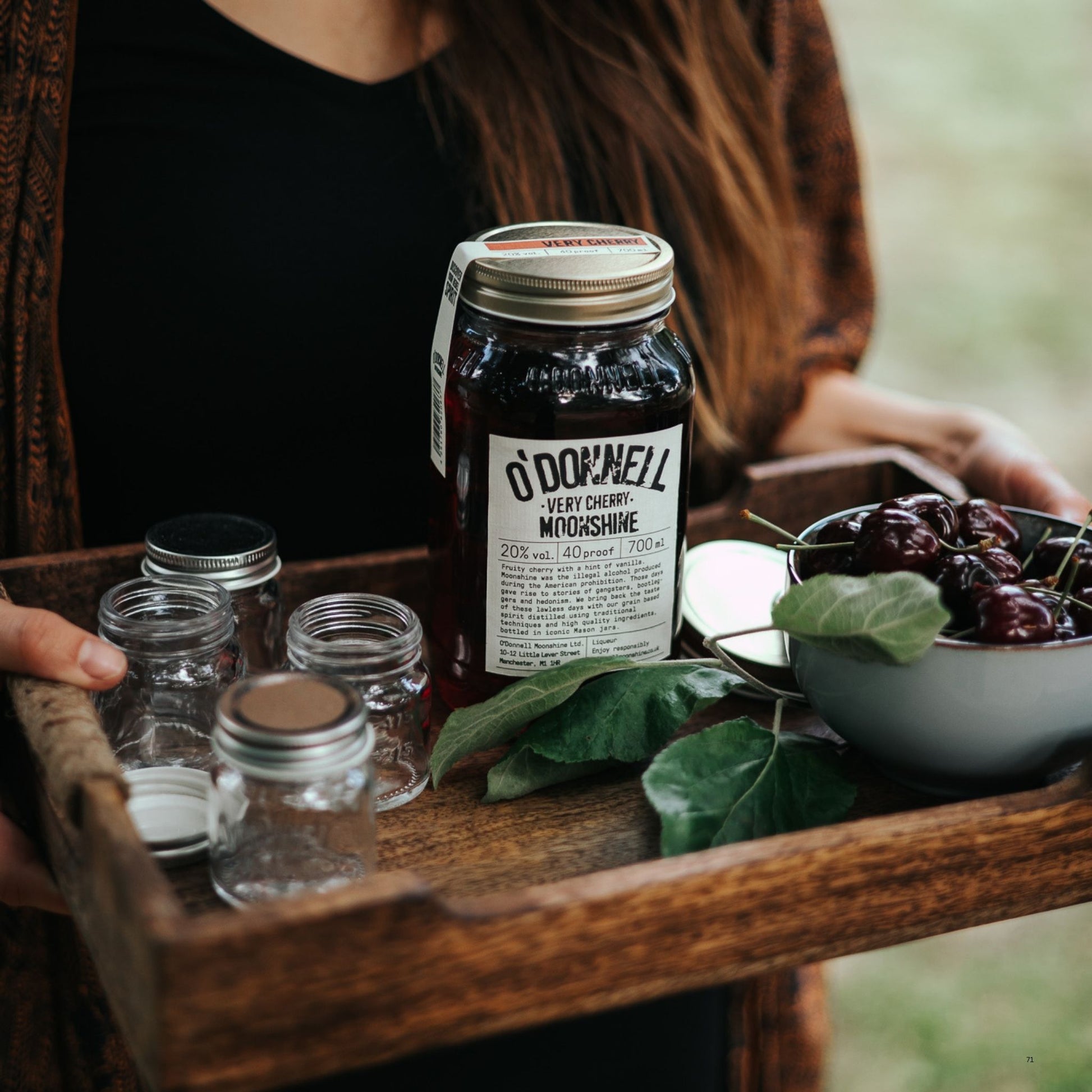 A wooden tray holds a jar of O'Donnell Very Cherry Moonshine alongside several small empty glass jars, green leaves, and a bowl of dark cherries. A person in a black top and brown jacket is partially visible holding the tray in an outdoor setting.
