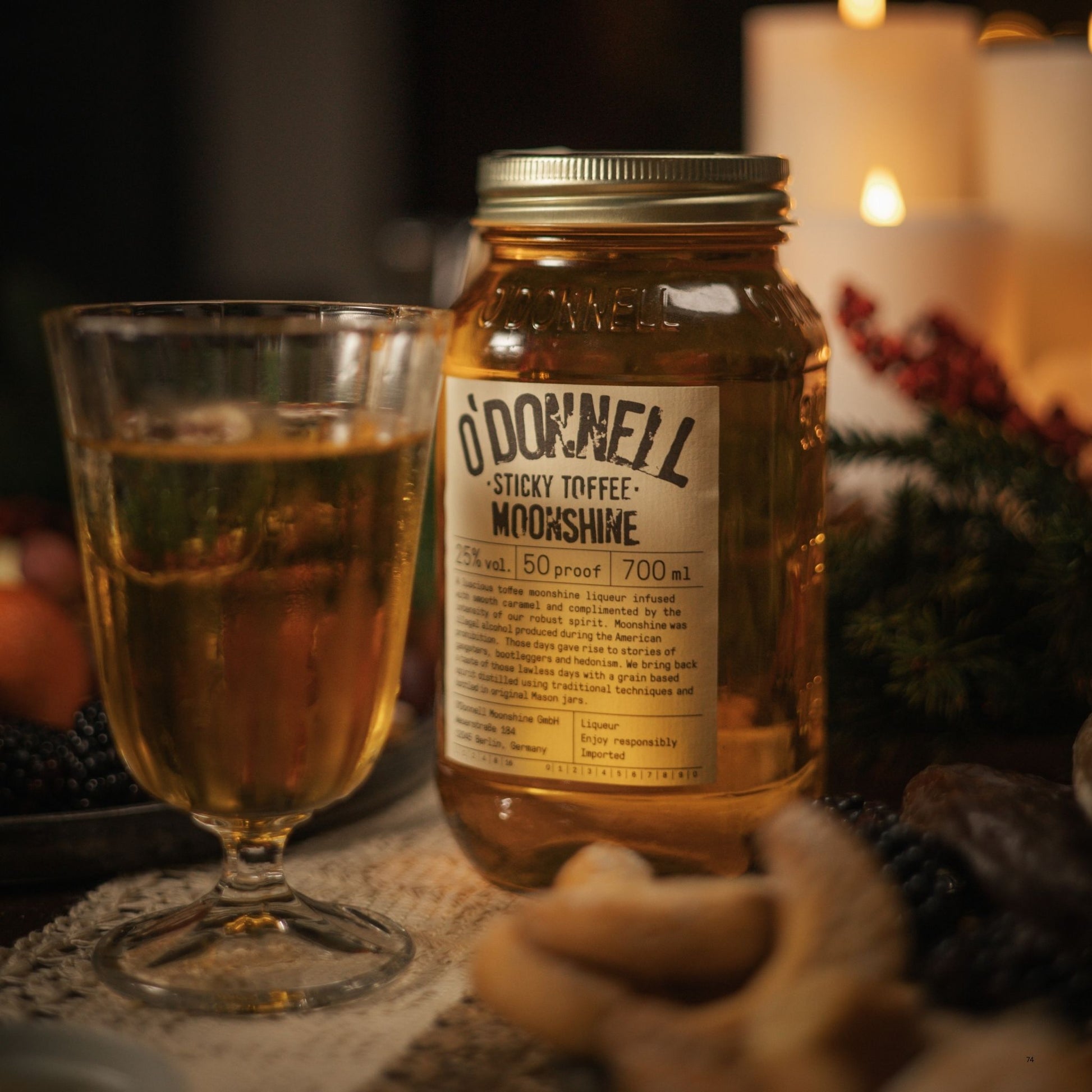 A glass of amber liquid sits beside a large clear jar labeled 'O'Donnell Sticky Toffee Moonshine' on a rustic tabletop with warm candlelight in the background.