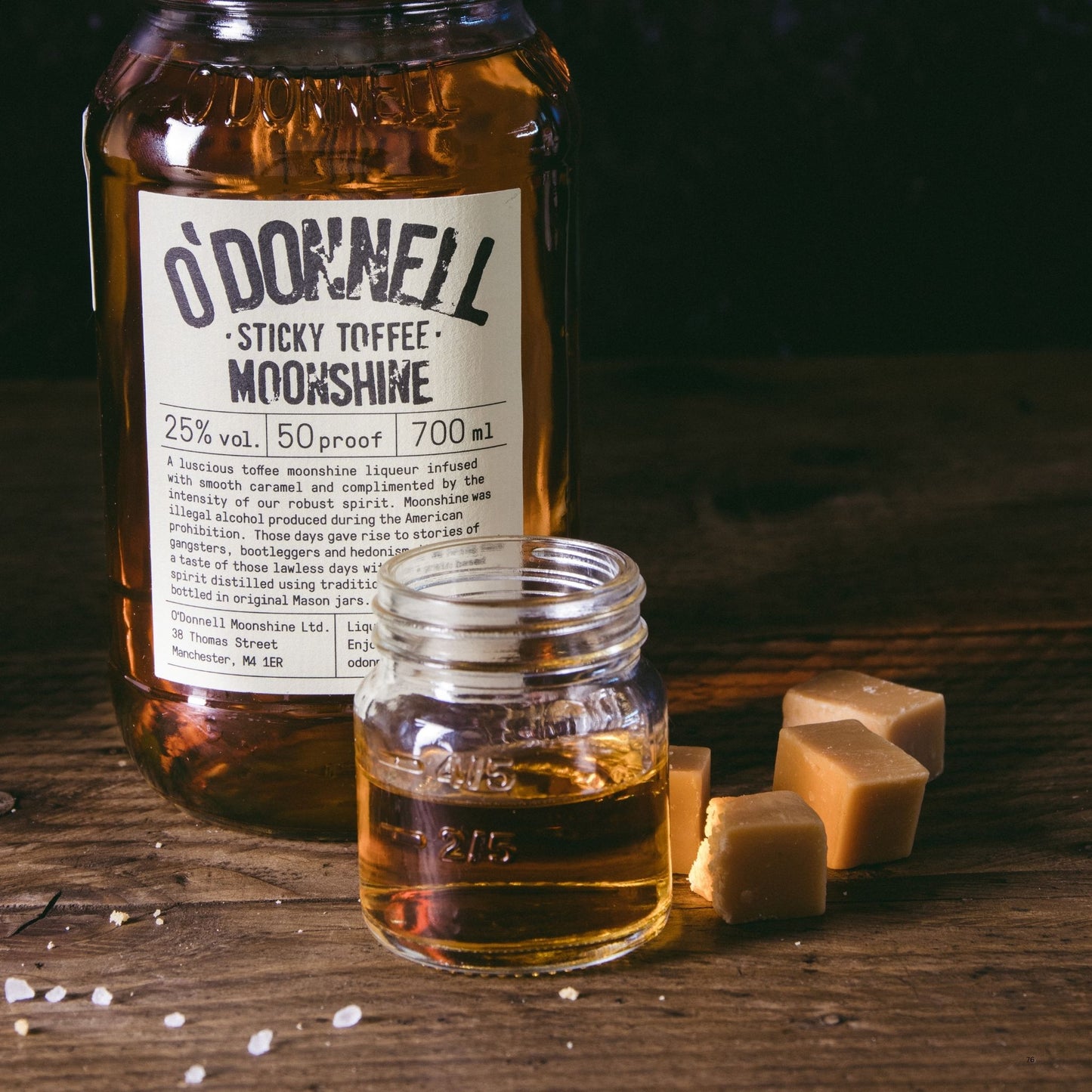 A large glass bottle of O'Donnell Sticky Toffee Moonshine stands on a wooden surface next to a smaller open glass jar filled with the amber liquid, with several square toffee cubes nearby.
