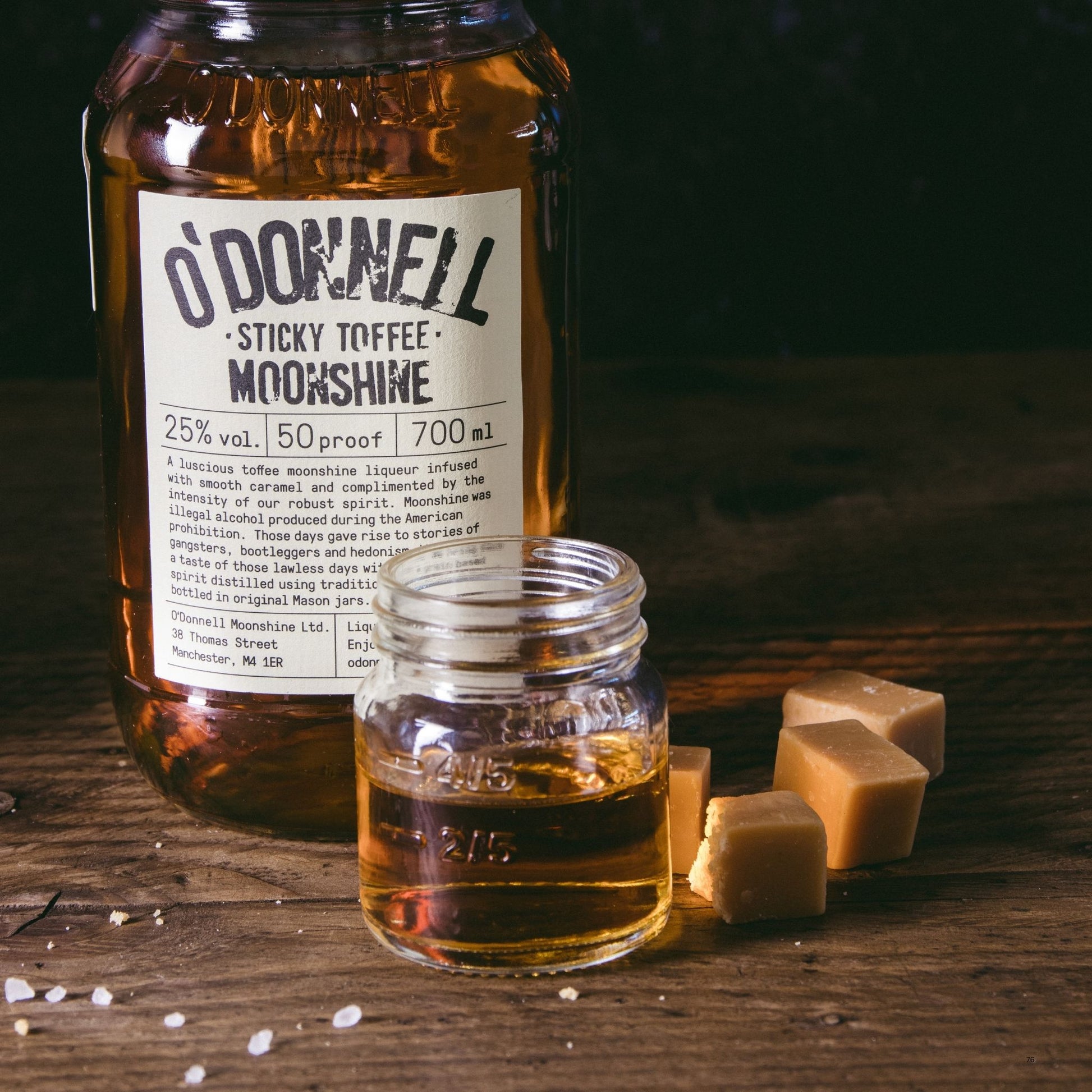 A large glass bottle of O'Donnell Sticky Toffee Moonshine stands on a wooden surface next to a smaller open glass jar filled with the amber liquid, with several square toffee cubes nearby.