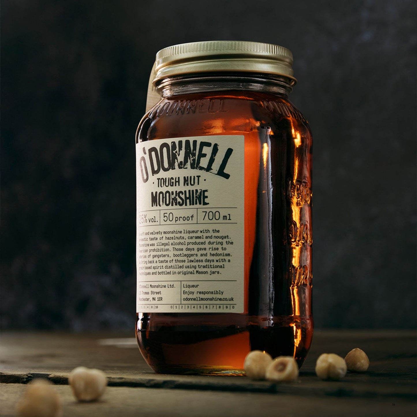 A glass jar of O'Donnell Tough Nut Moonshine with a gold metal lid and a cream-colored label, set on a dark rustic wooden surface with hazelnuts scattered around. The background is a dark, textured studio setting.