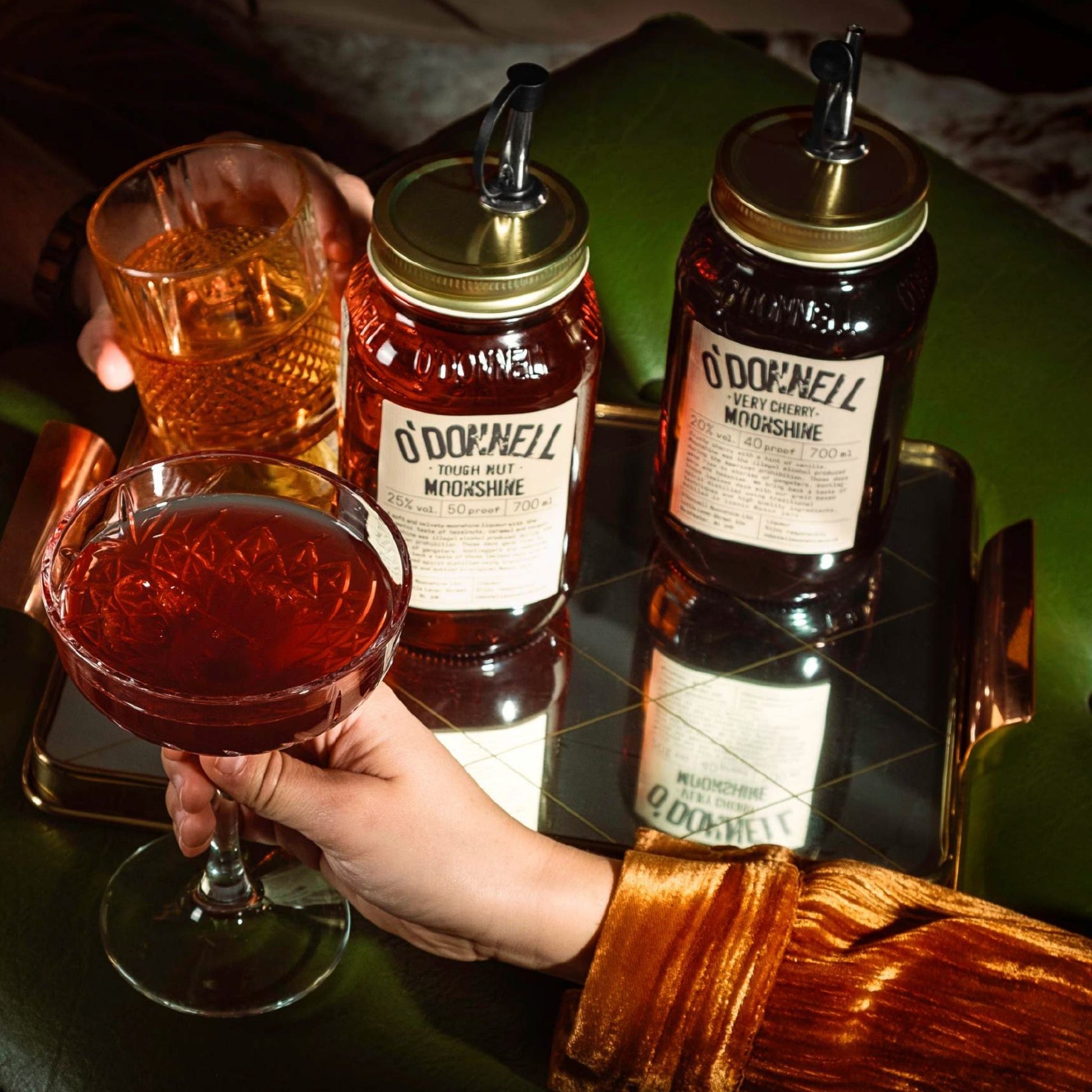Two glass bottles of O'Donnell liqueur with gold lids and black straws sit on a reflective tray beside a coupe glass filled with amber liquid and a hand holding it. A person's hand and gold-veiled sleeve are visible in a dim, moody setting.
