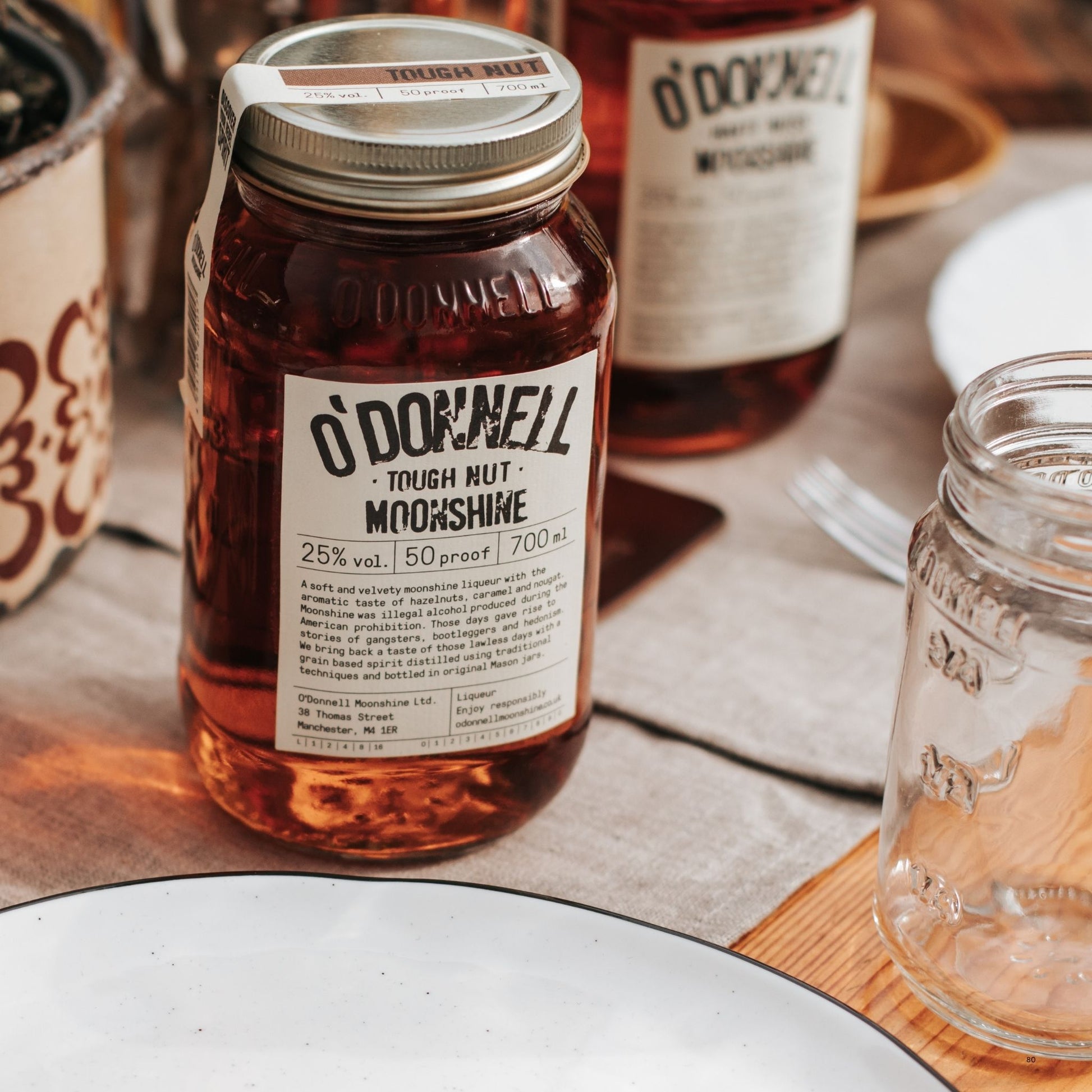 A glass mason jar filled with amber liquor, labeled O'Donnell Tough Nut Moonshine 25% vol. 50 proof, 700 ml, set on a wooden table with plates and another bottle in the background.