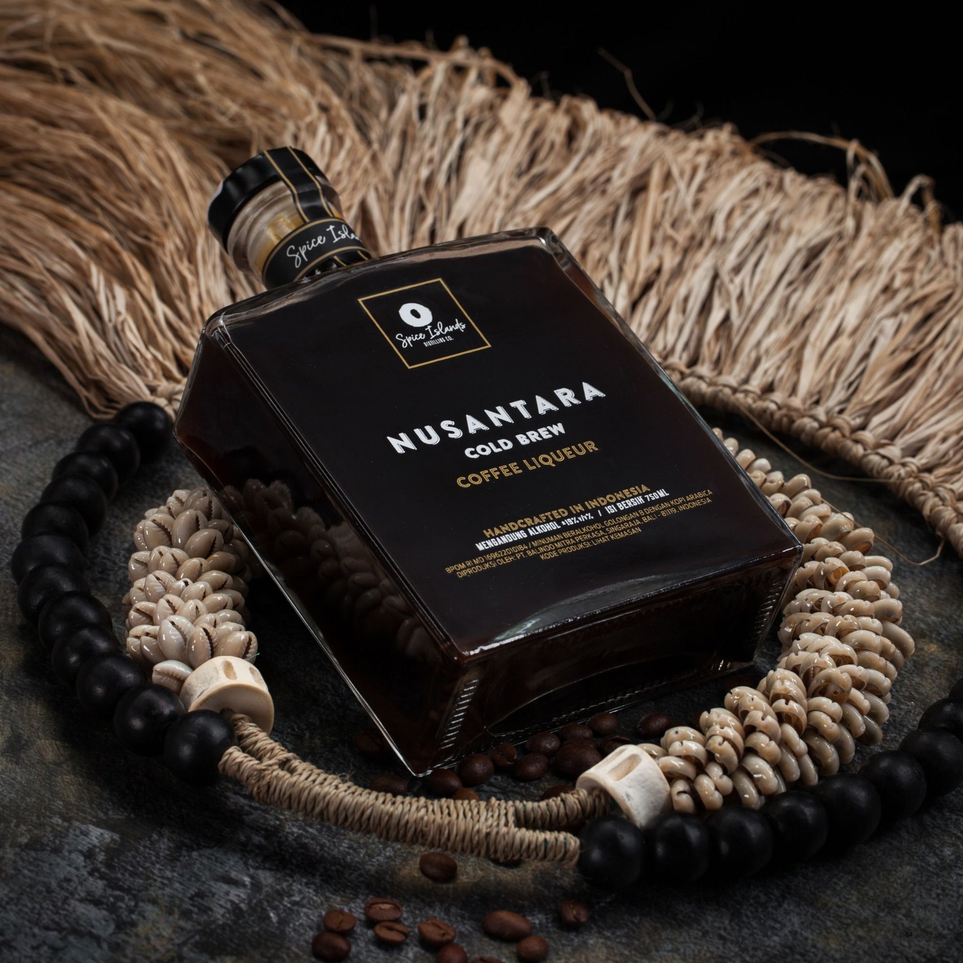A bottle of Nusantara Cold Brew Coffee Liqueur, styled with coffee beans, a woven straw mat, and a beaded necklace on a dark textured surface.
