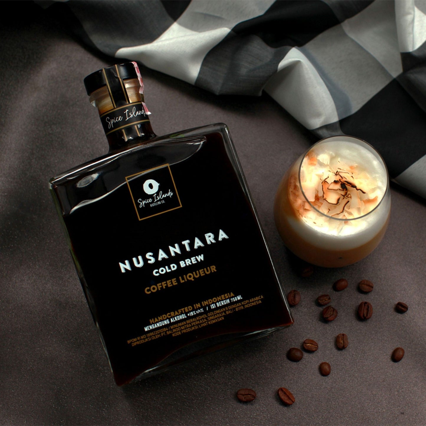 A bottle of Nusantara Cold Brew Coffee Liqueur by Spice Islands is styled on a dark textured surface next to a glass of iced coffee with whipped cream and coffee beans, with a black and white checkered cloth in the background.