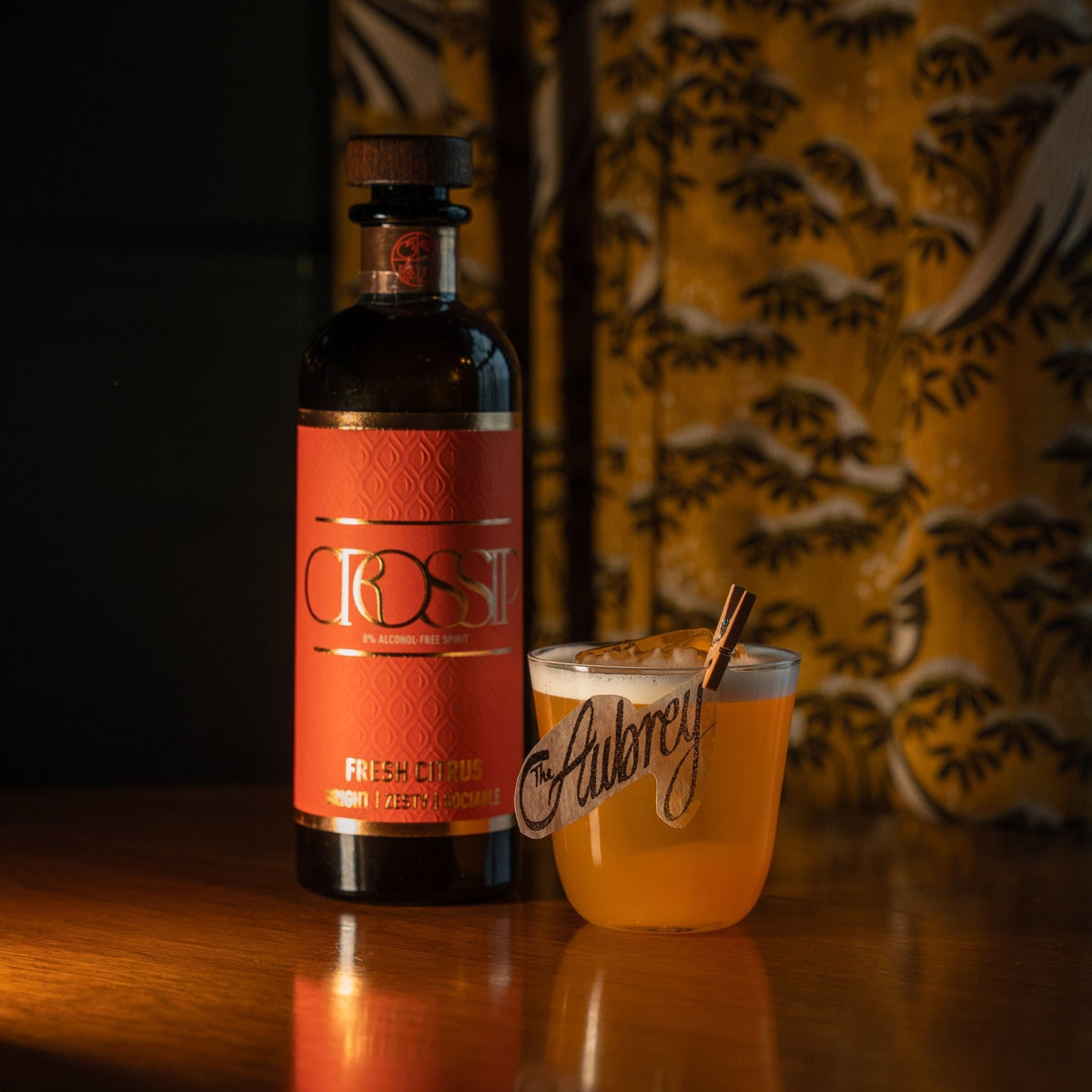 A bottle of CROSSIP Fresh Citrus non-alcoholic citrus liqueur stands on a wooden table next to a frothy orange cocktail in a glass with the CROSSIP logo, set against a warm, patterned yellow and brown background.