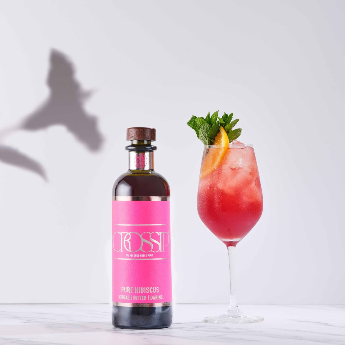 A bottle of CROSSIP Pure Hibiscus next to a pink hibiscus cocktail in a stemmed glass, garnished with mint and orange slice, on a light marble surface with a white background and a bird shadow.