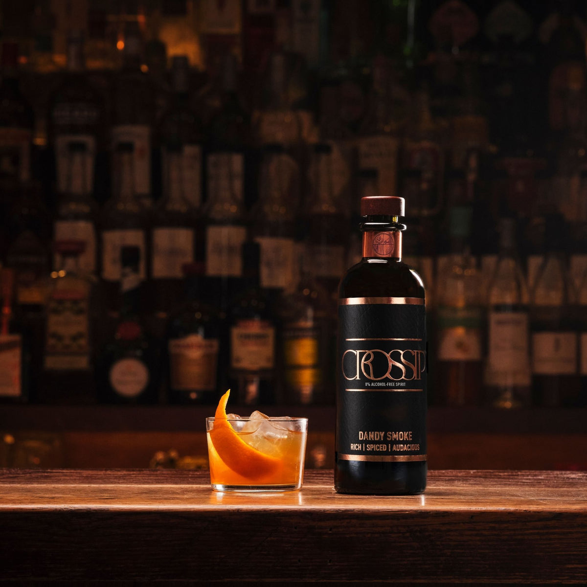 A bottle of Crossip Spirits Dandy Smoke cocktail mix stands on a wooden bar counter next to a glass filled with an orange-colored drink and an orange peel garnish, with a blurred bar shelf of bottles in the background.
